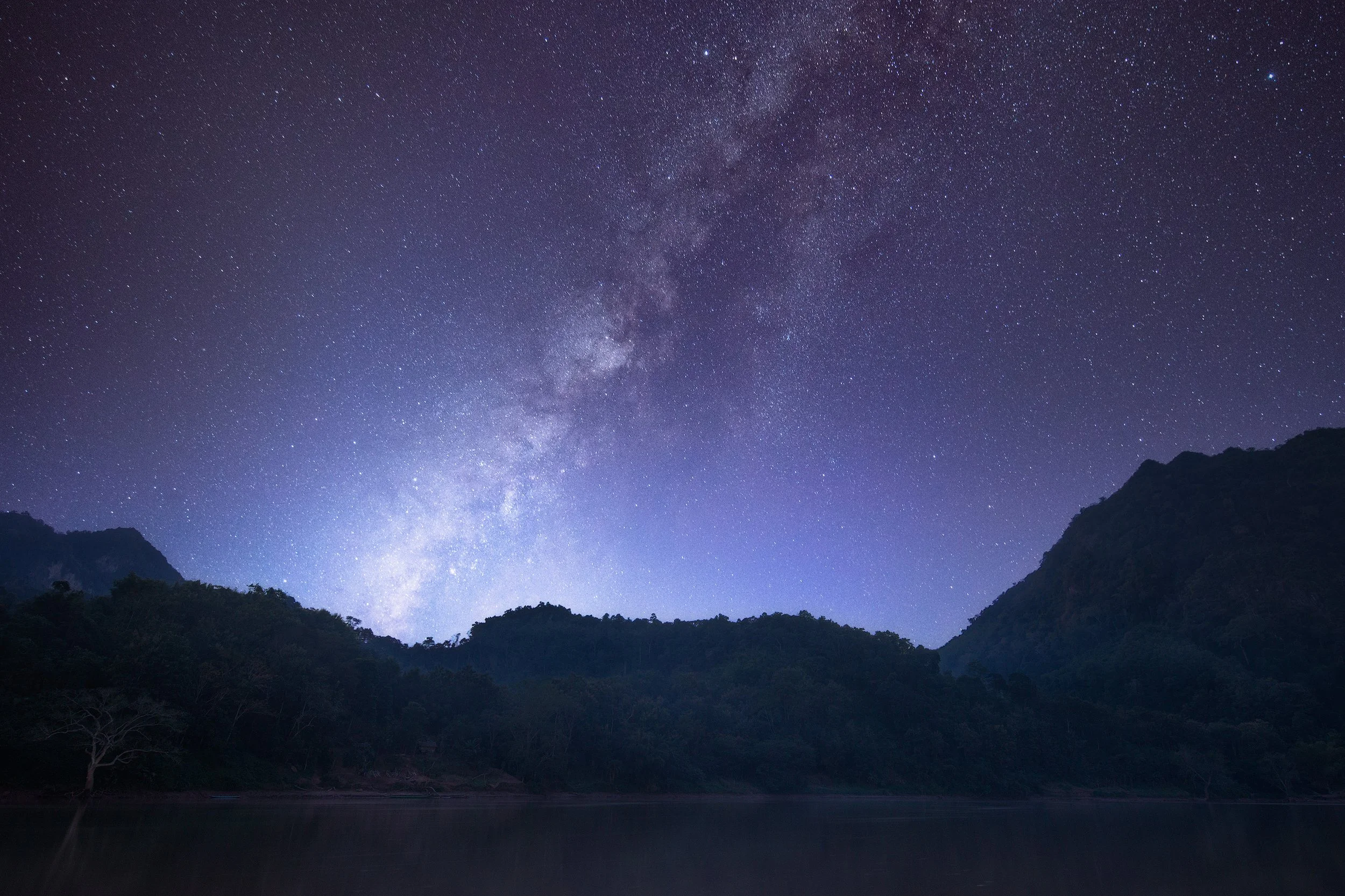 Night sky filled with stars and the Milky Way galaxy, with dark mountain silhouettes and a calm lake in the foreground.