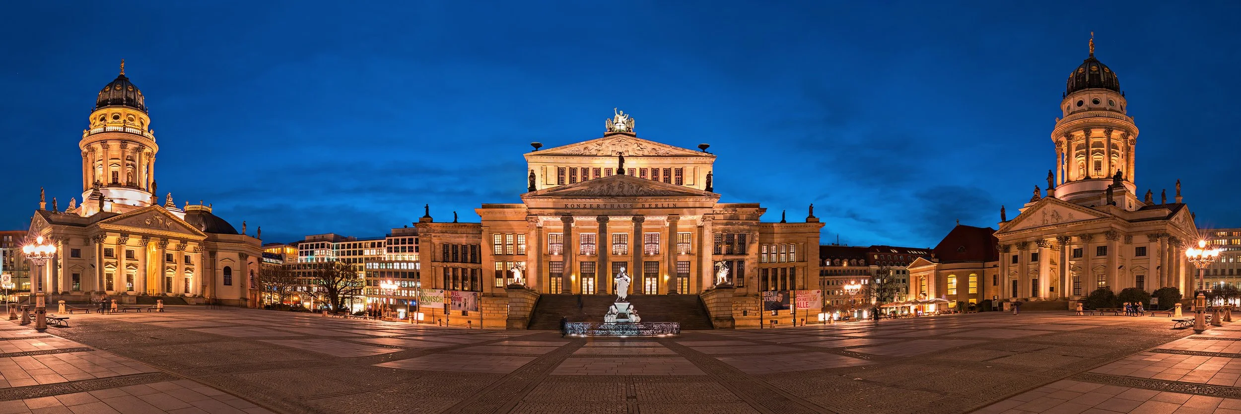 Night view of Konzerthaus Berlin, with illuminated historic buildings and statues in front, under a deep blue sky.