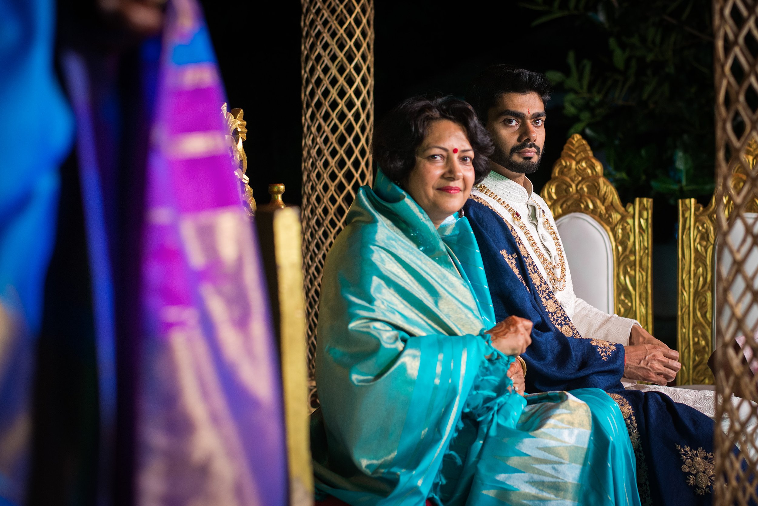An elderly woman and a young man sitting on decorated chairs at an event, with colorful fabrics and golden ornamentation around them, outdoors at night.