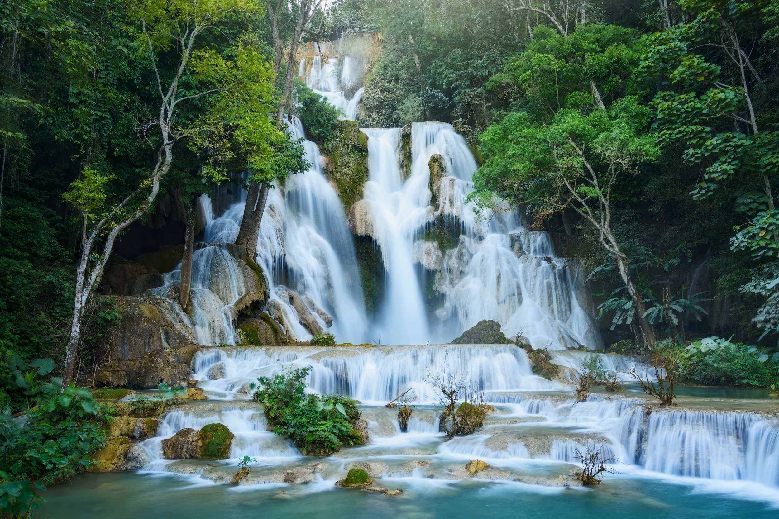 Multi-tiered waterfall cascading over rocks surrounded by lush green trees in a tropical forest. Laos.