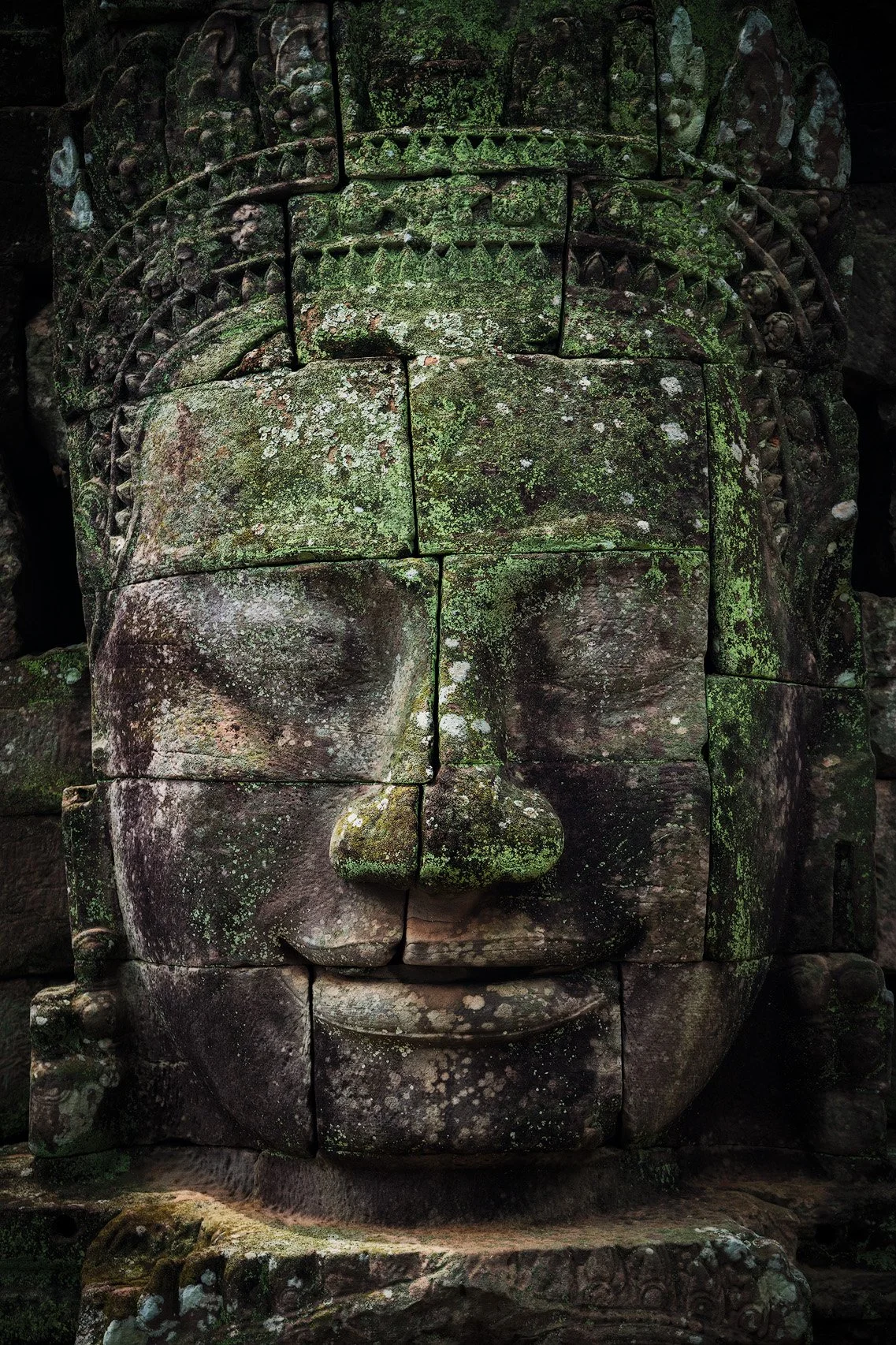 Close-up of an ancient stone carving of a face with moss and lichen growing on it, featuring closed eyes, a pronounced nose, and a slight smile, likely part of a larger structure or statue.