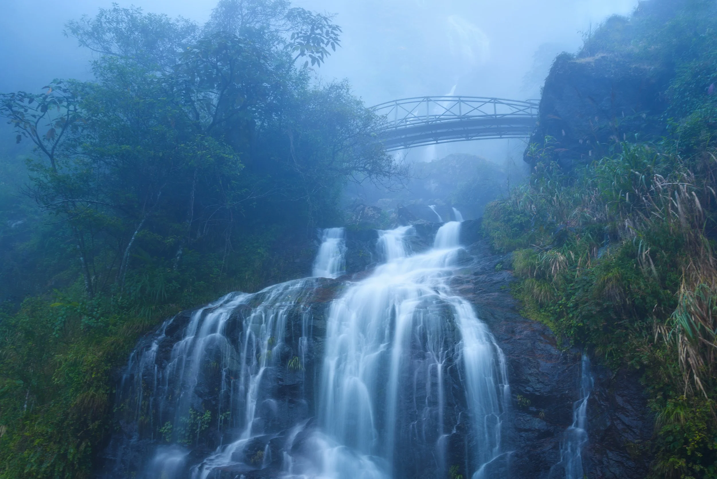 A misty waterfall flowing down a rocky cliff surrounded by lush green foliage, with a metal bridge visible in the background. Vietnam, Sapa.