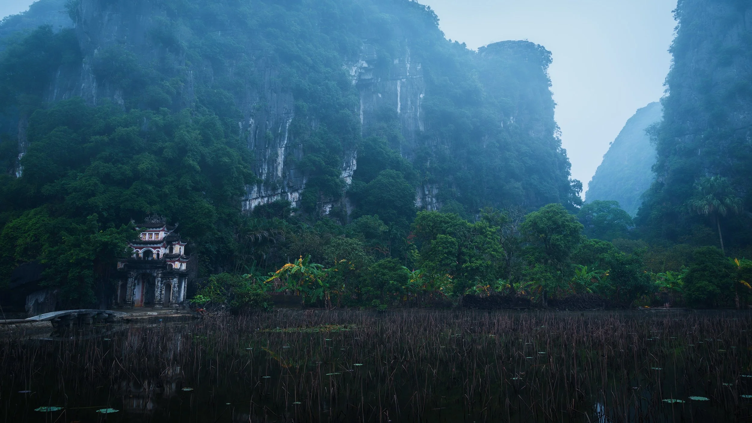A lush green valley surrounded by tall, mist-covered mountains with a small, ornate building at the base of one mountain.