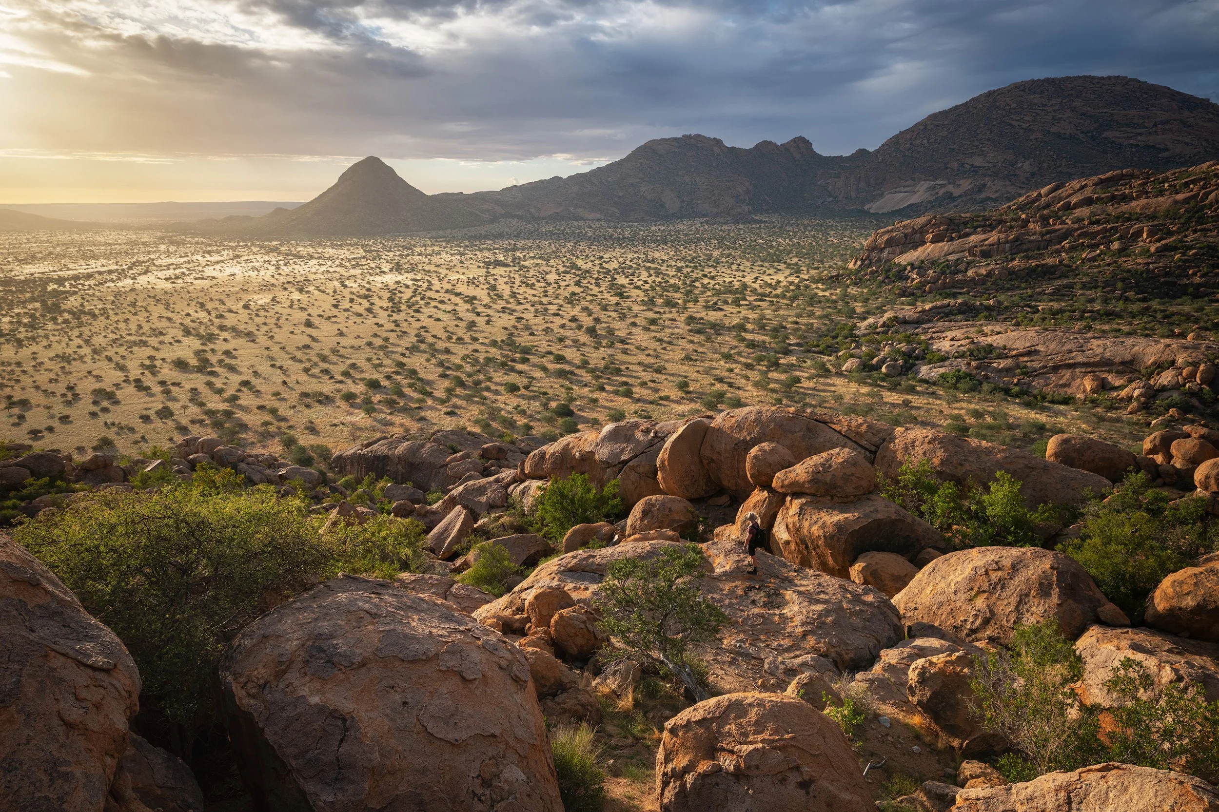 A desert landscape with large rocks and sparse green bushes in the foreground, mountain ranges in the distance, and a partly cloudy sky with the sun setting or rising. Erongo, Namibia