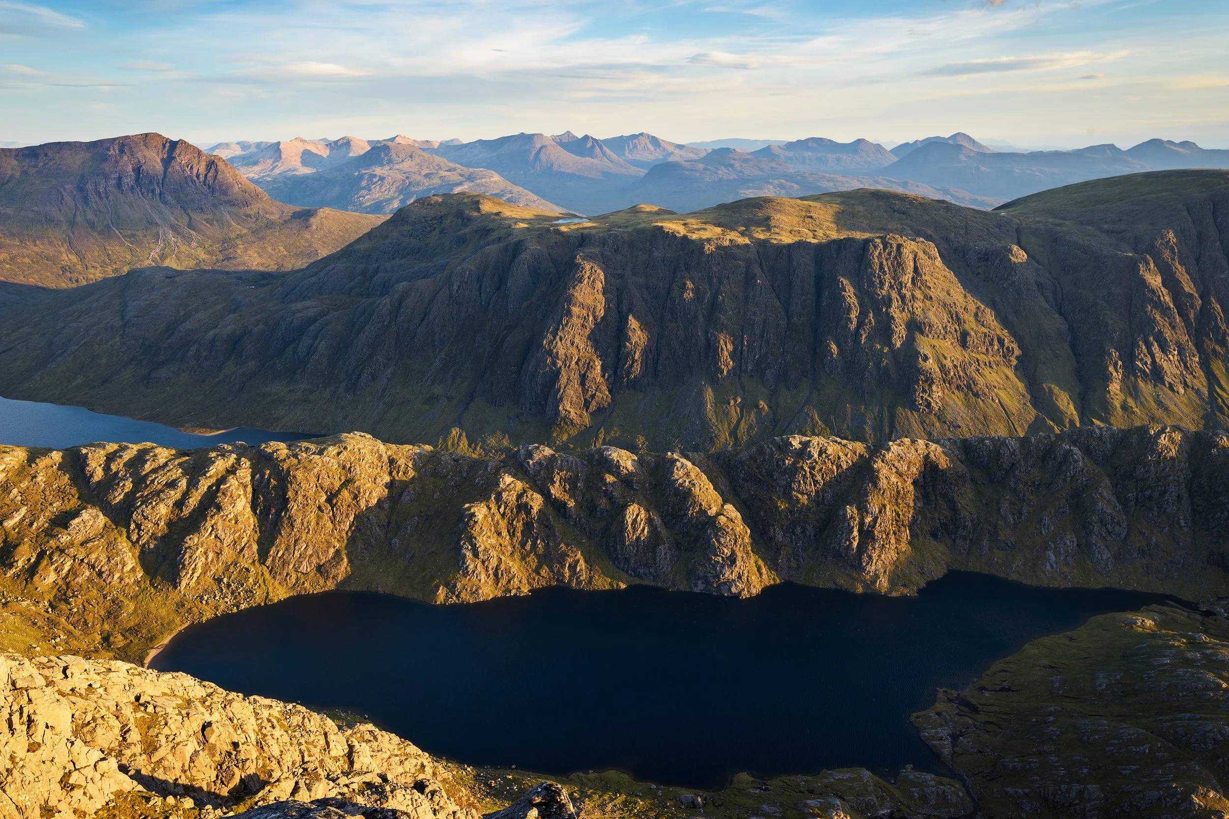 Mountain landscape with a large lake in the foreground, rugged peaks, and hills under a partly cloudy sky.