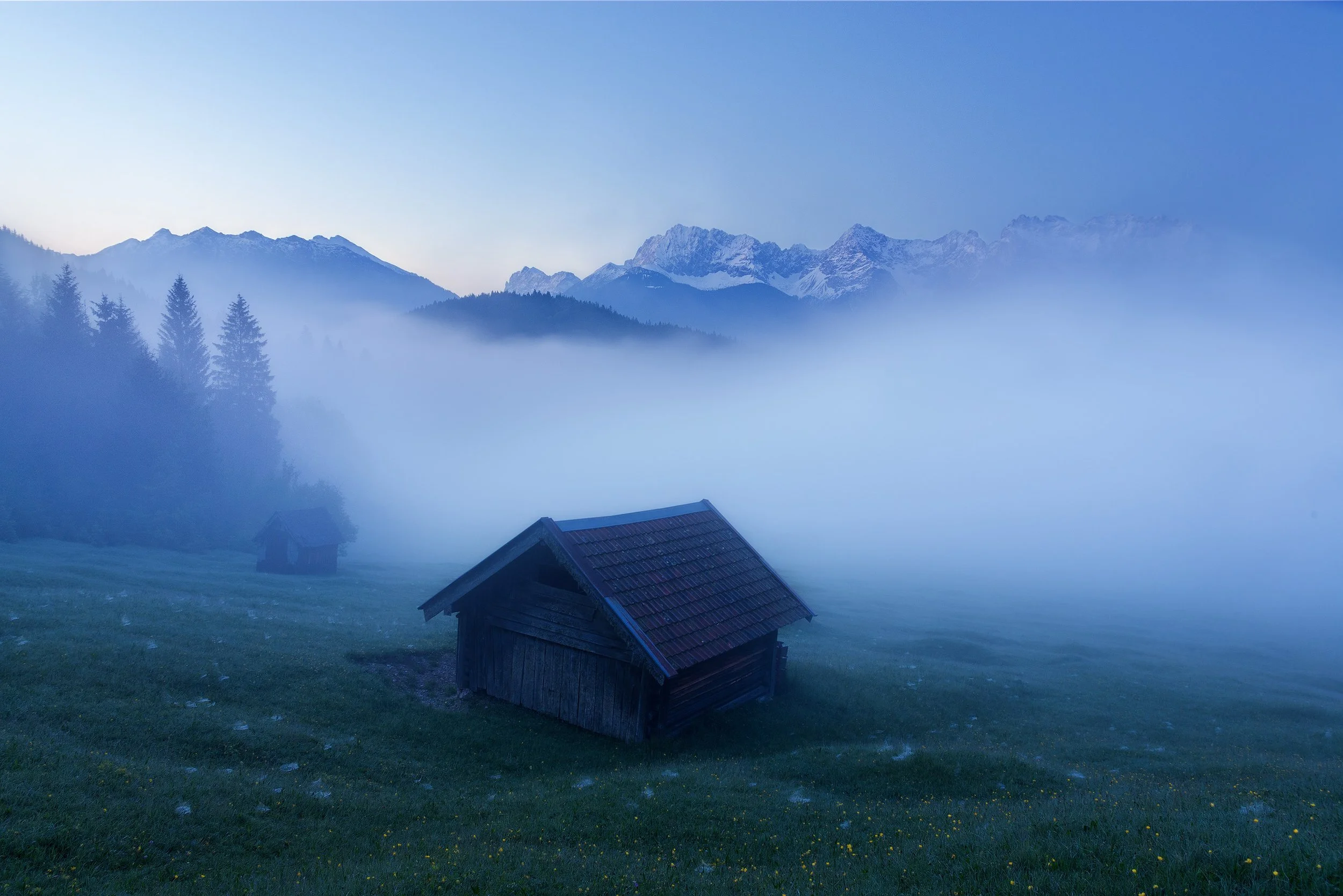 Gerold See in Bavaria, a mountain landscape with fog, trees, and two small wooden cabins on a grassy slope