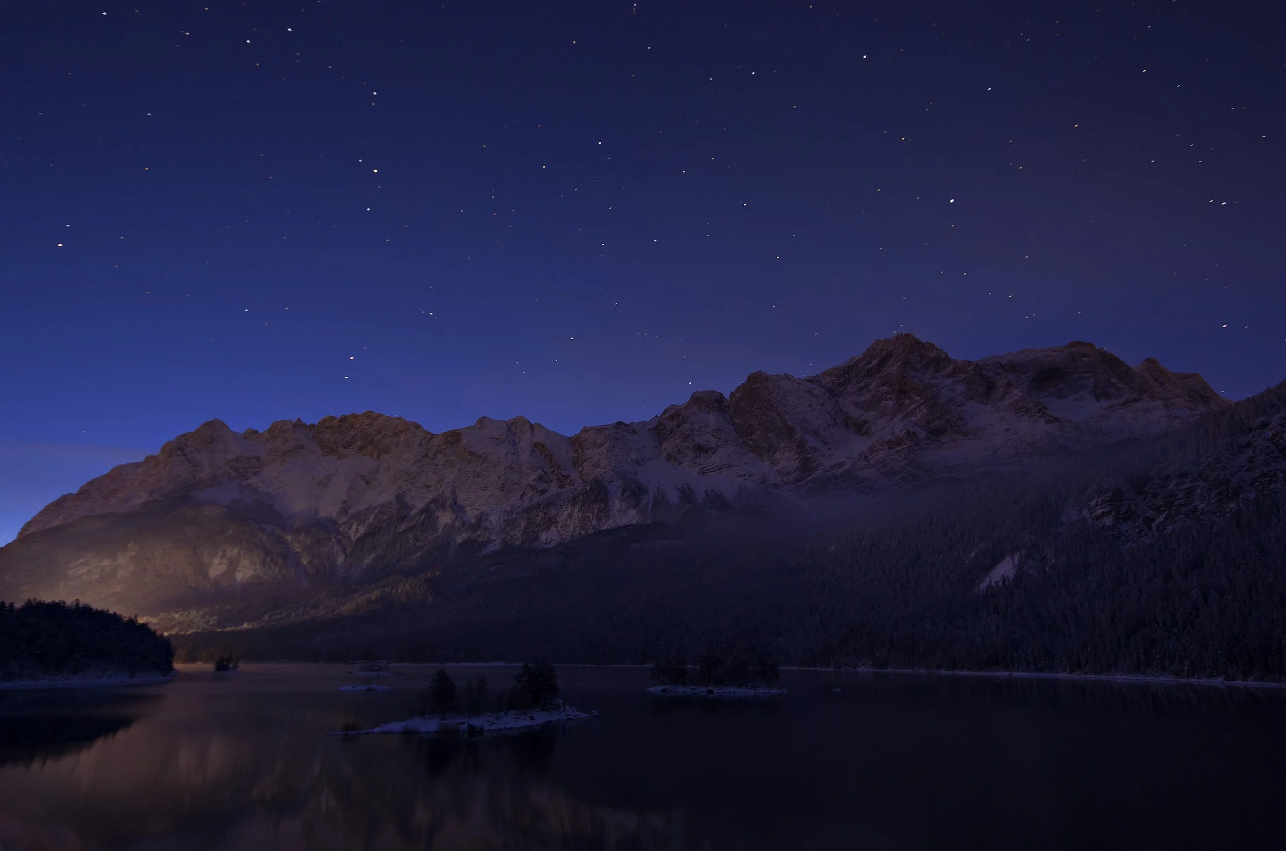 Nighttime landscape of snow-covered mountain range overlooking the calm Eibsee in Bavaria with small islands, under a starry sky.