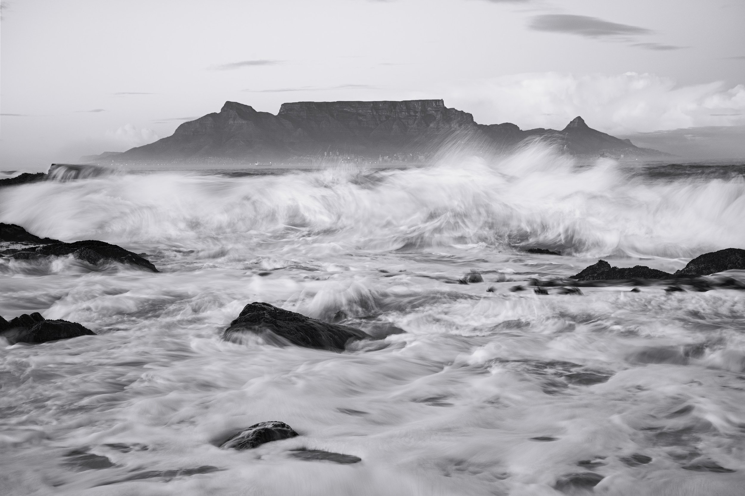 Black and white photo of ocean waves crashing on rocks with Table Mountain in the background. cape town South Africa.