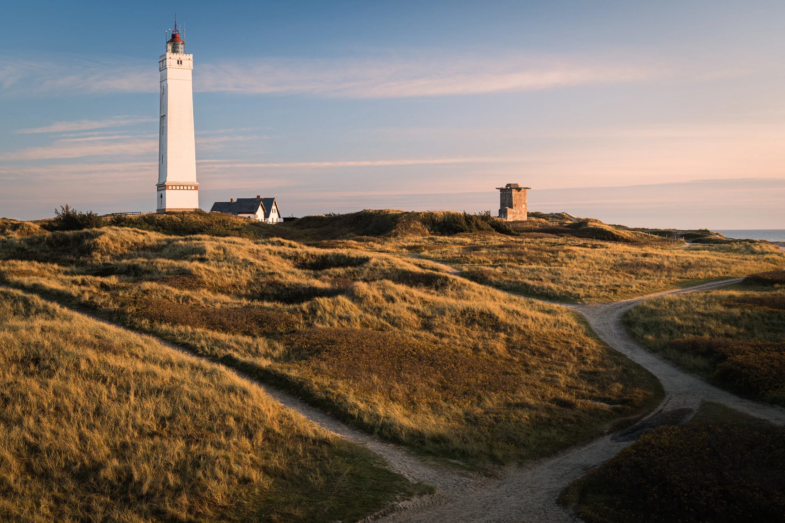 A tall white lighthouse on a grassy hill with dirt paths and a small building nearby, under a partly cloudy sky during sunset.