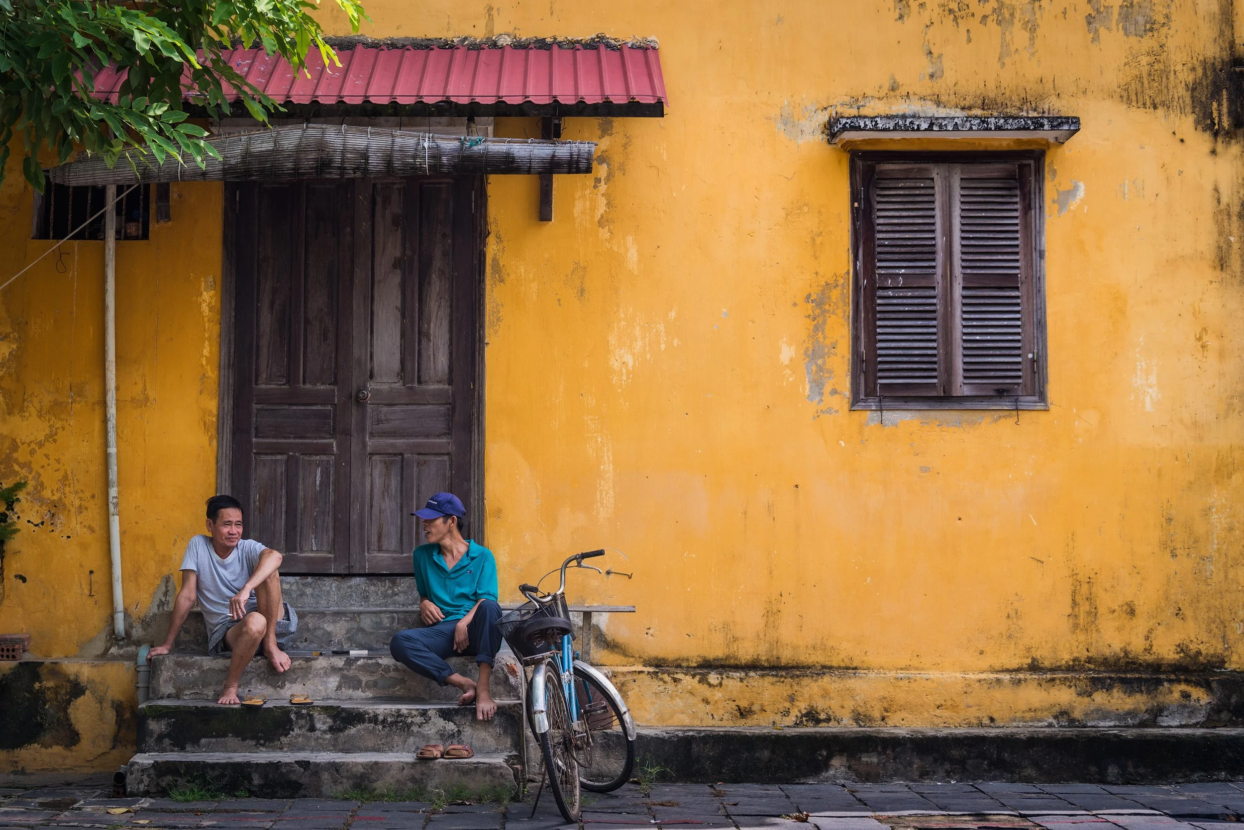 Two people sitting on stairs outside a yellow wall with a wooden door and window, a bicycle leaning against the stairs, and some greenery in the top left corner.