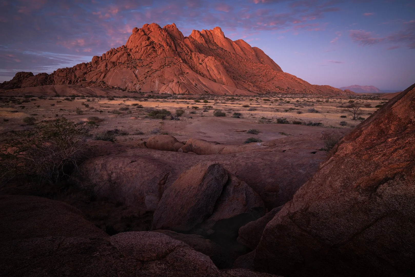 A spitzkoppe landscape in Namibia with large reddish rock formations under a pinkish-purple sky at sunset.
