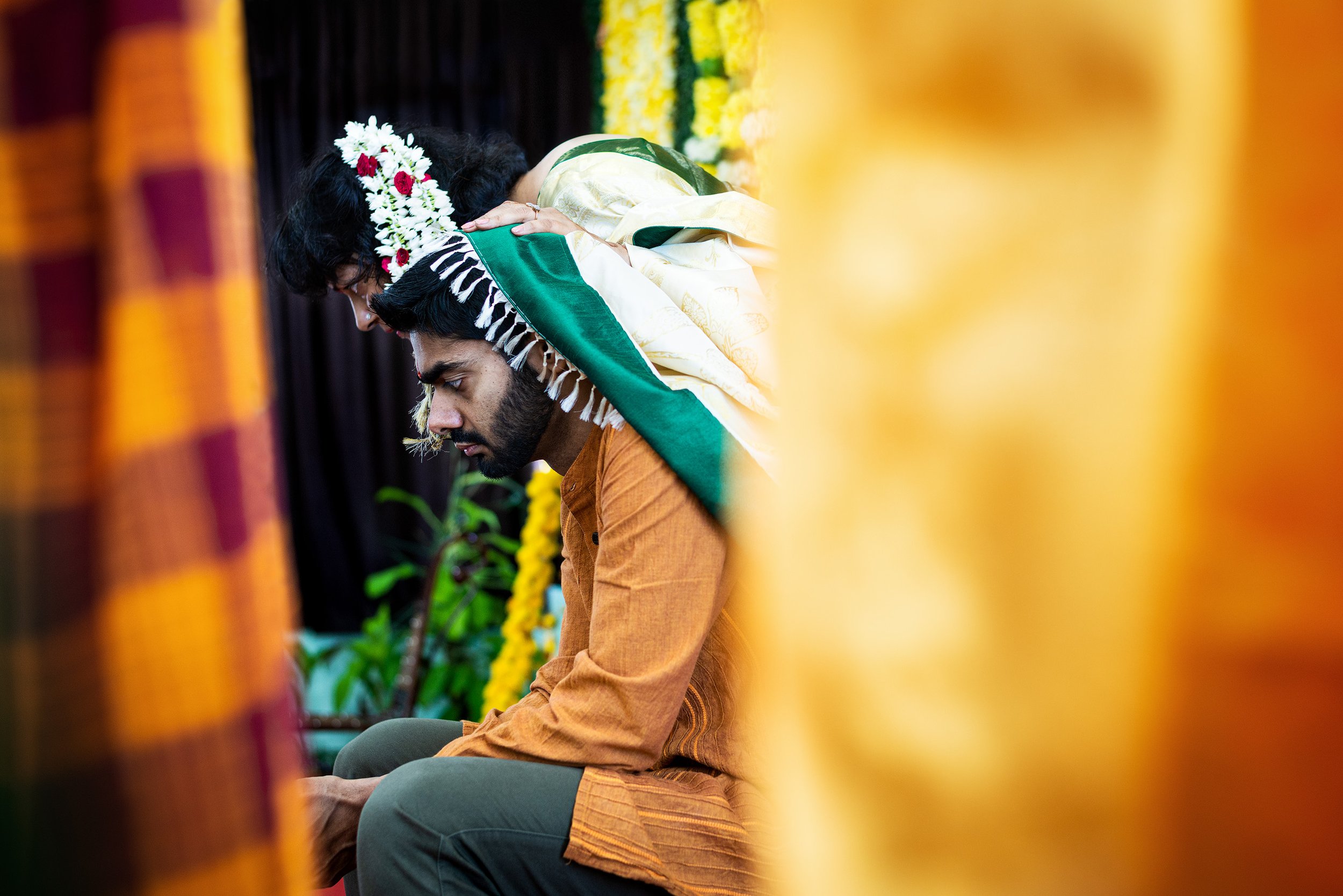 A man sitting with his head bowed, wearing traditional clothing, with a woman leaning on his shoulder, in a decorated setting with yellow flowers and floral arrangements in the background.