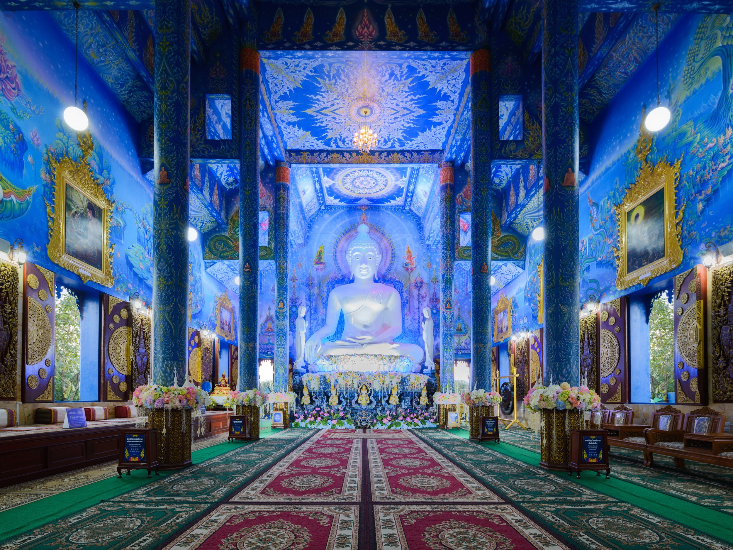 Interior of a Buddhist temple featuring a large illuminated white Buddha statue surrounded by colorful floral arrangements and intricate blue and gold decorations on the walls and ceiling.