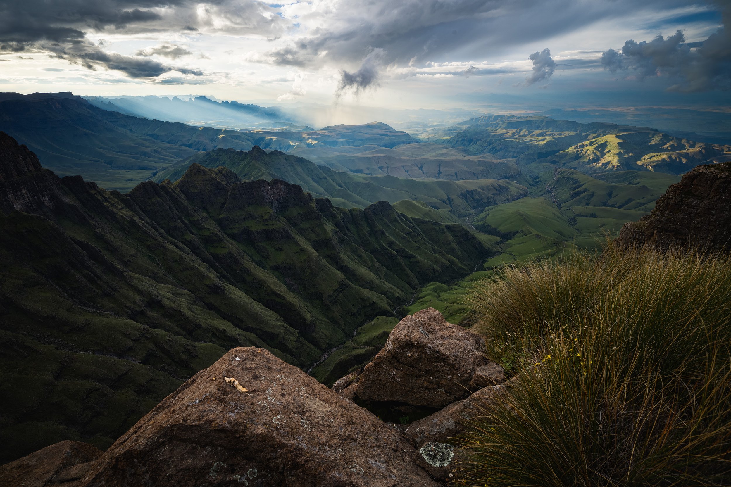 A vast green mountainous landscape under a cloudy sky, with rugged ridges, valleys, and patches of sunlight breaking through the clouds. Drakensberg Mountains, South Africa.