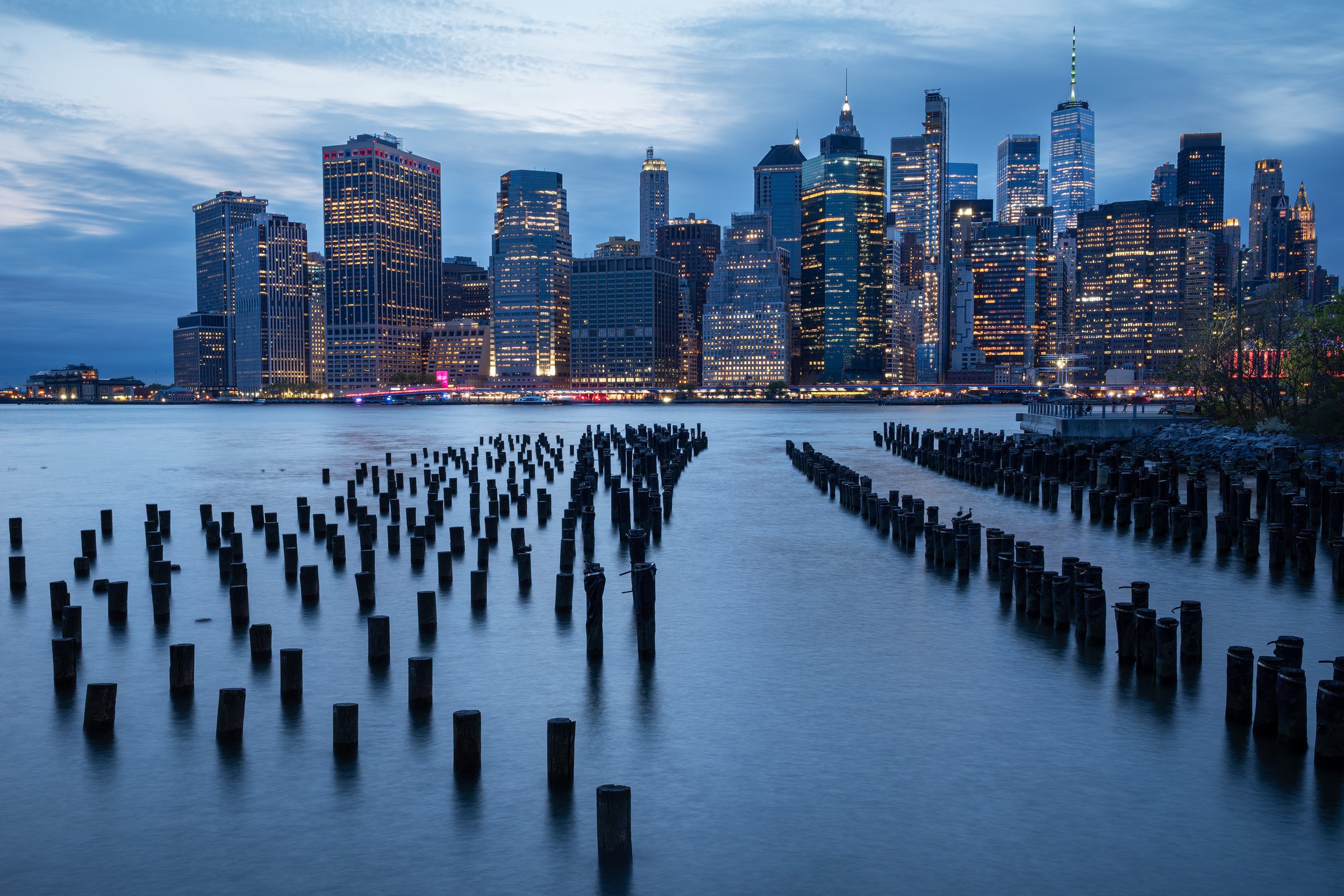 City skyline at dusk with illuminated skyscrapers across the water, with wooden pilings in the foreground.