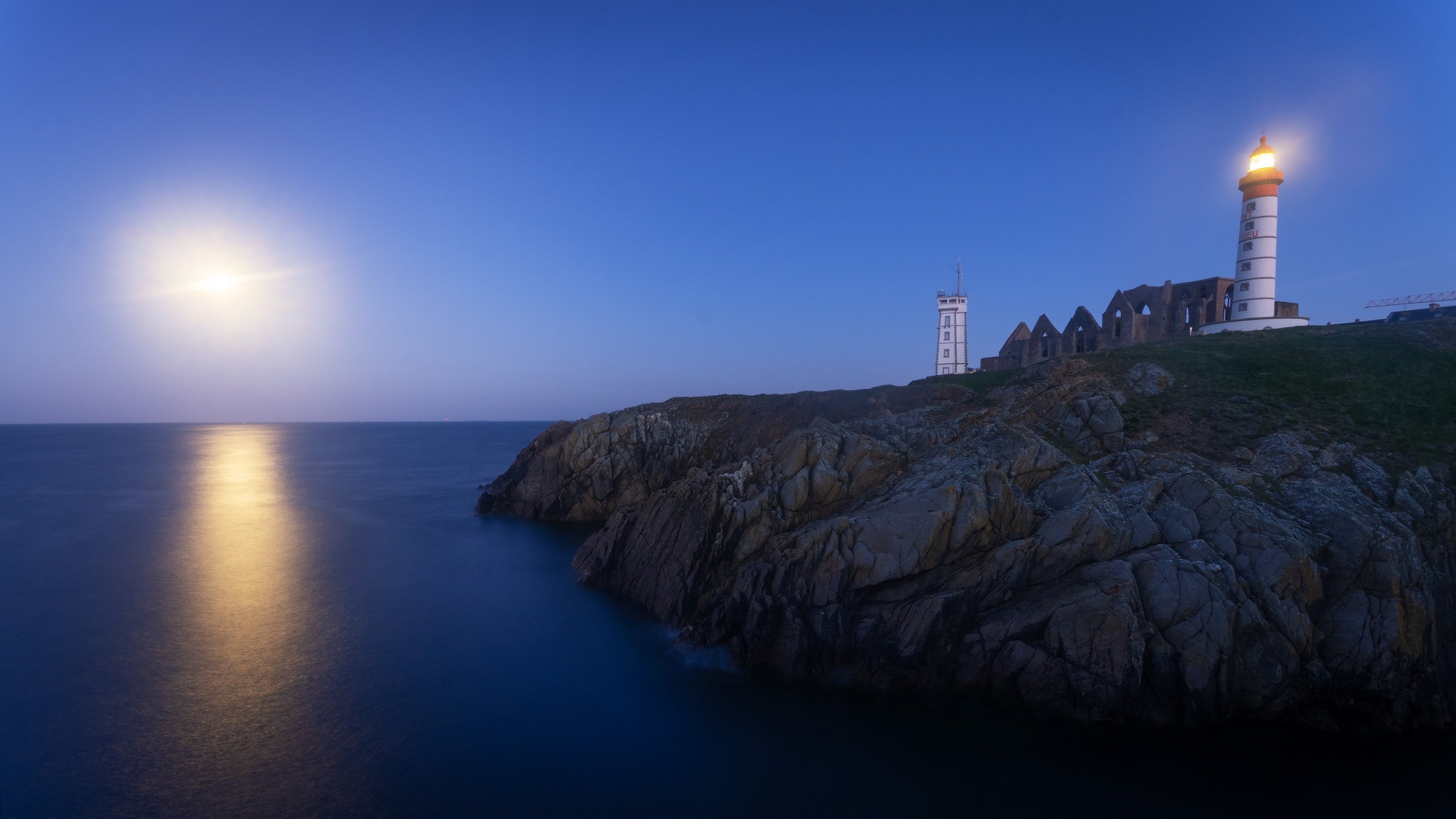A lighthouse on a rocky coastline under a moonlit sky, with the moon reflecting on the calm sea.