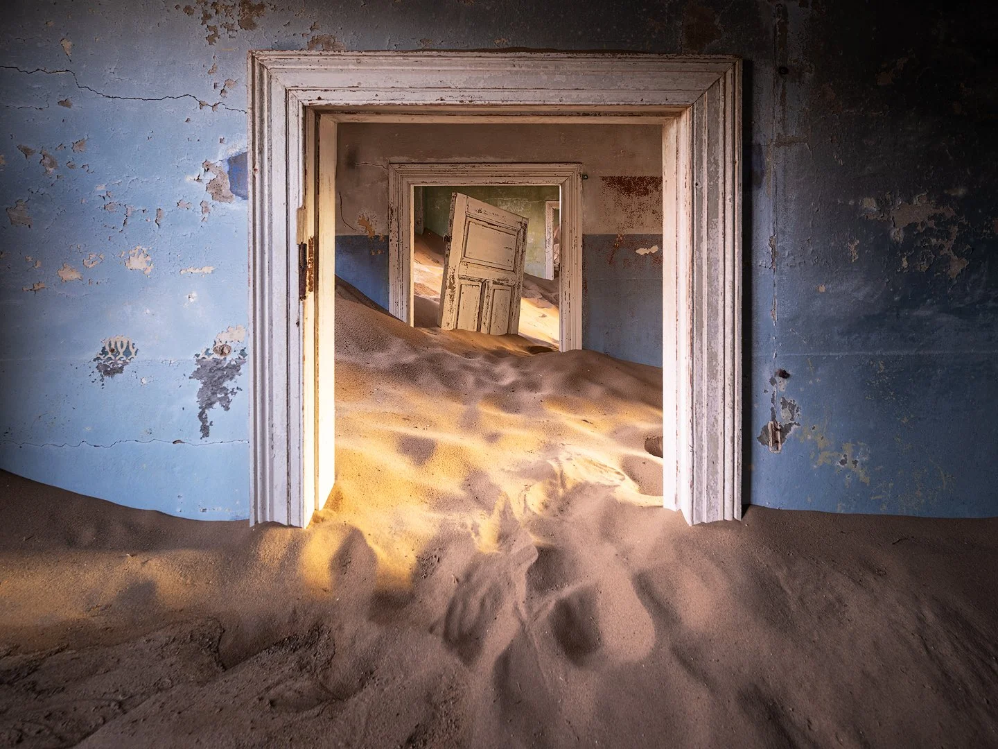 An abandoned house in Kolmanskoppe, Namibia, with multiple doorways, filled with sand that has blown inside, creating dunes along the floors.
