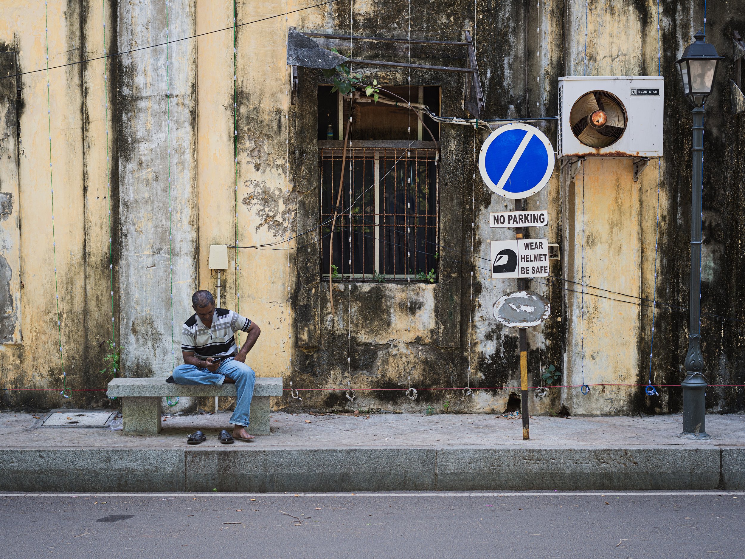 A man sitting on a bench on a sidewalk in front of an old, weathered yellow wall with a barred window, surrounded by various street signs including a 'No Parking' sign and a sign instructing to wear a helmet and be safe. The man appears to be using a