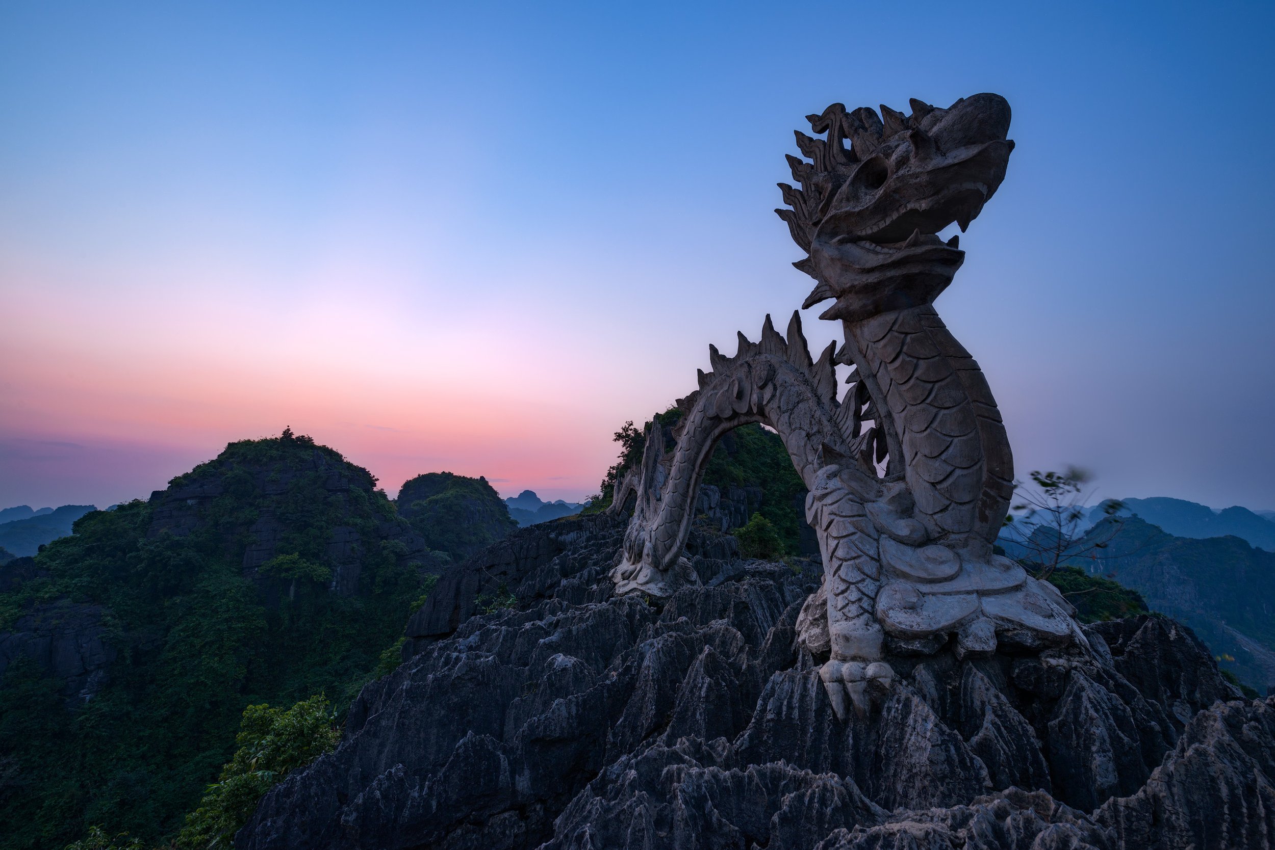 Stone dragon sculpture on rocky landscape with distant mountains and a colorful sunset sky. Vietnam, Tam Coc.