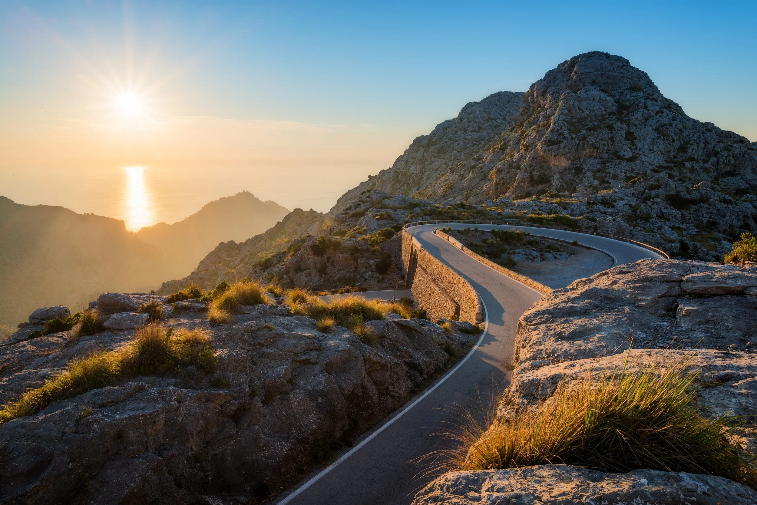 A winding mountain road at sunset with rocky terrain and sparse vegetation, overlooking the ocean in the distance. Sa Calibre, Mallorca.