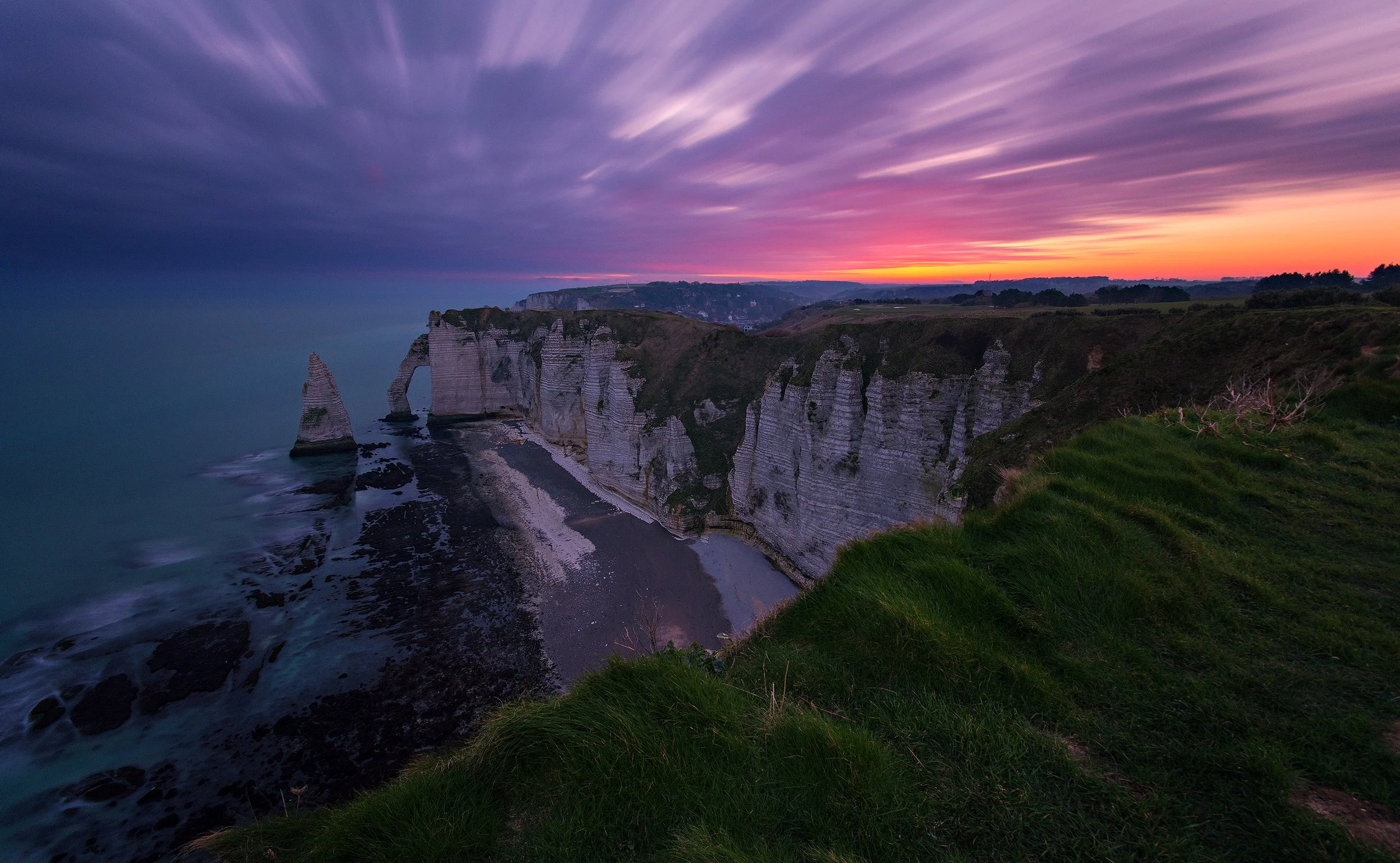 Cliffs along a shoreline at sunset with purple, pink, and orange sky, and grassy foreground.