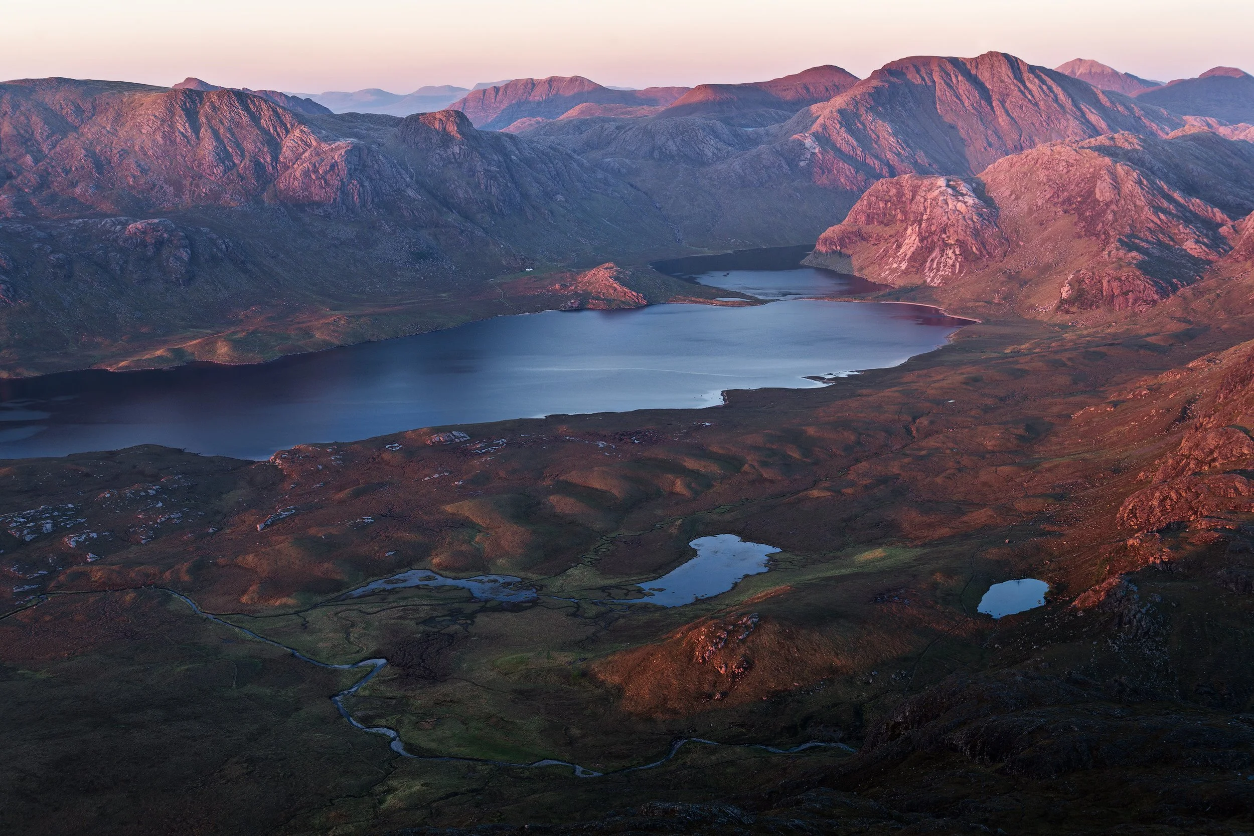 Scenic view of mountains with pinkish hues at sunset, featuring a large lake in the valley and smaller lakes nearby, with rugged terrain and a winding stream.