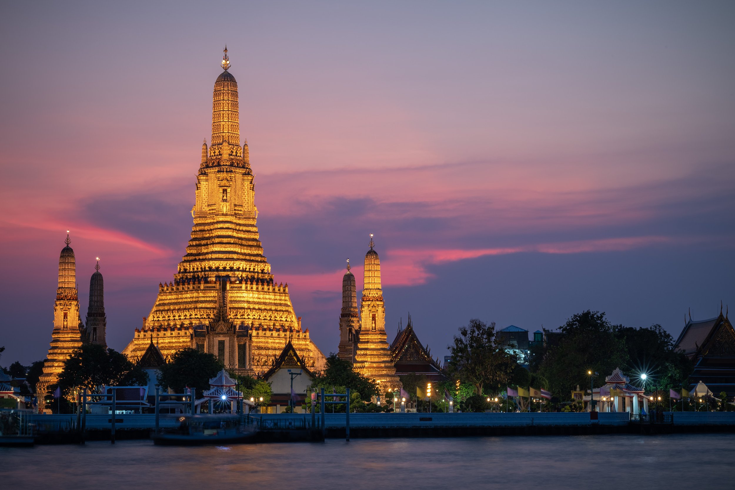 Nighttime view of Wat Arun temple illuminated with golden lights, with a pink and purple sky in the background and a river in the foreground.