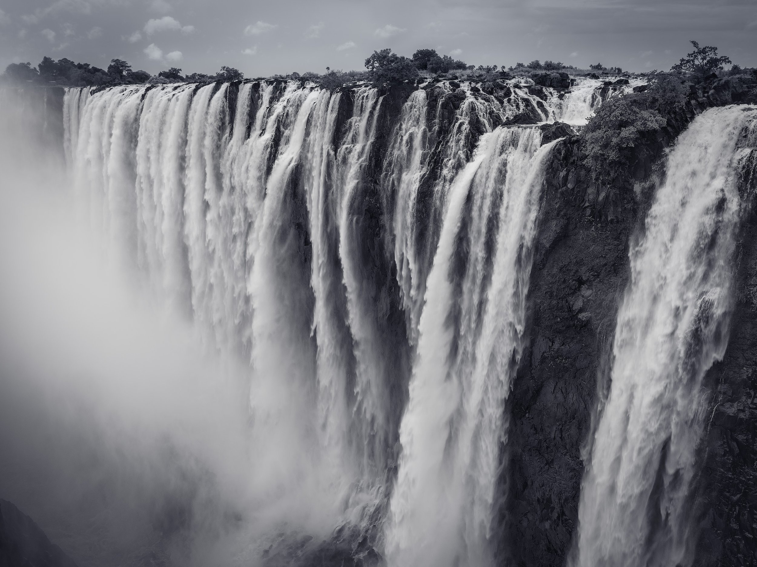 A large waterfall with multiple cascades flowing over rocky cliffs amidst mist and cloudy sky.