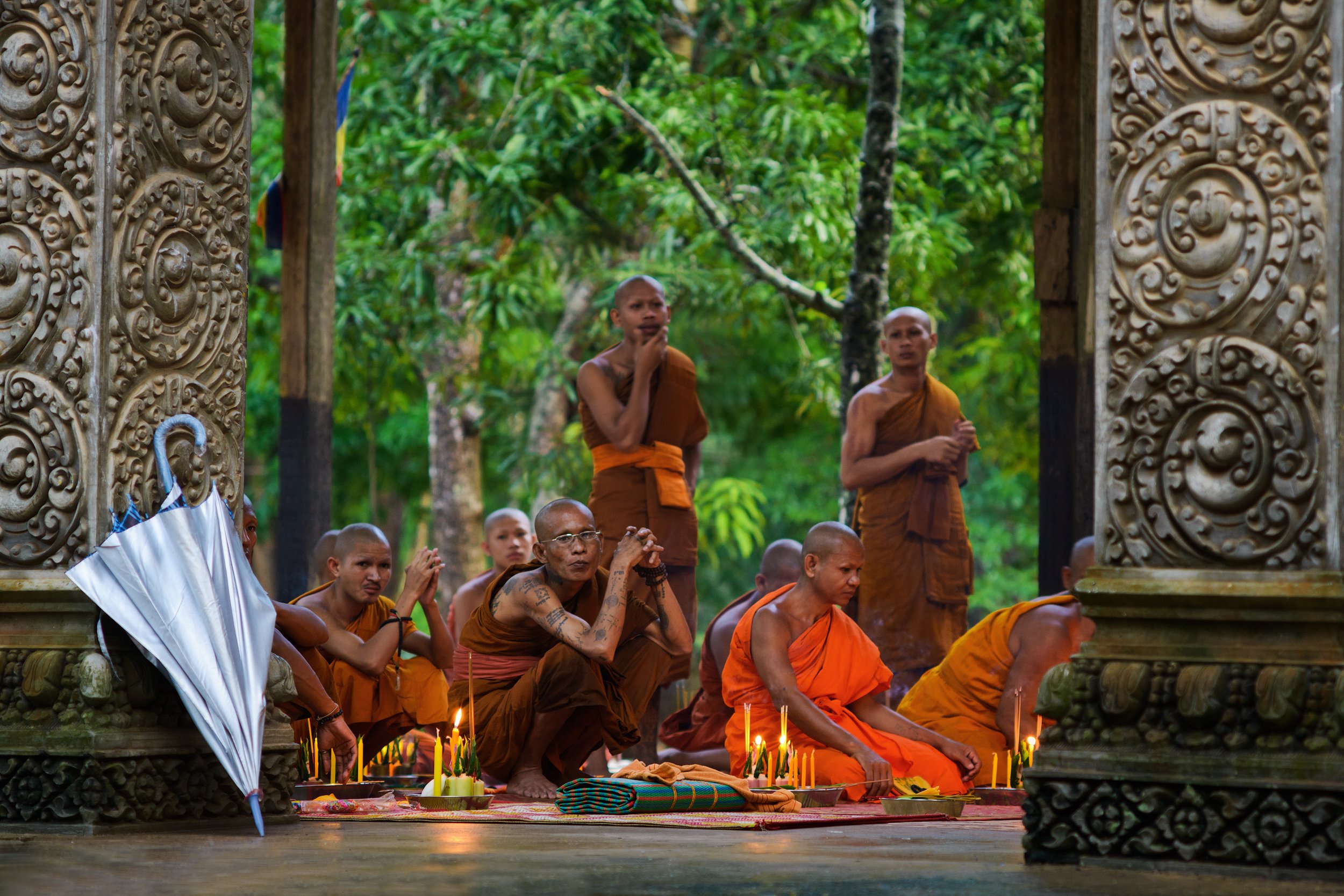 Buddhist monks and laypeople participating in a prayer or ceremony at a temple, with candles and offerings, set against a lush green forest.