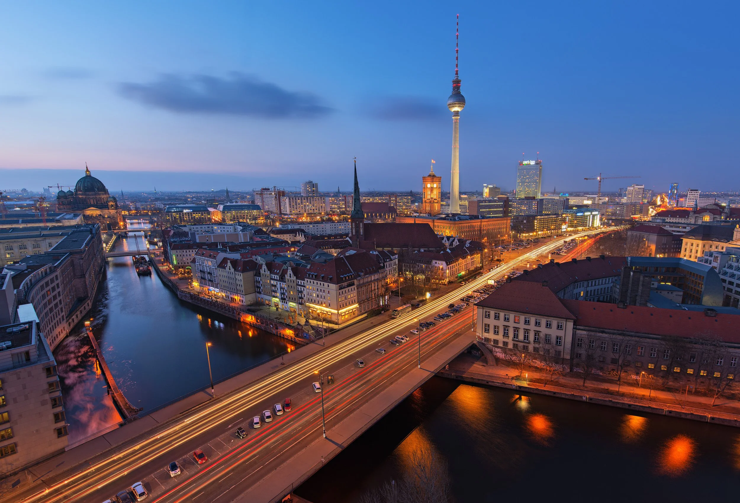 A city skyline at dusk with a river running through the city, featuring illuminated buildings and a tall television tower.
