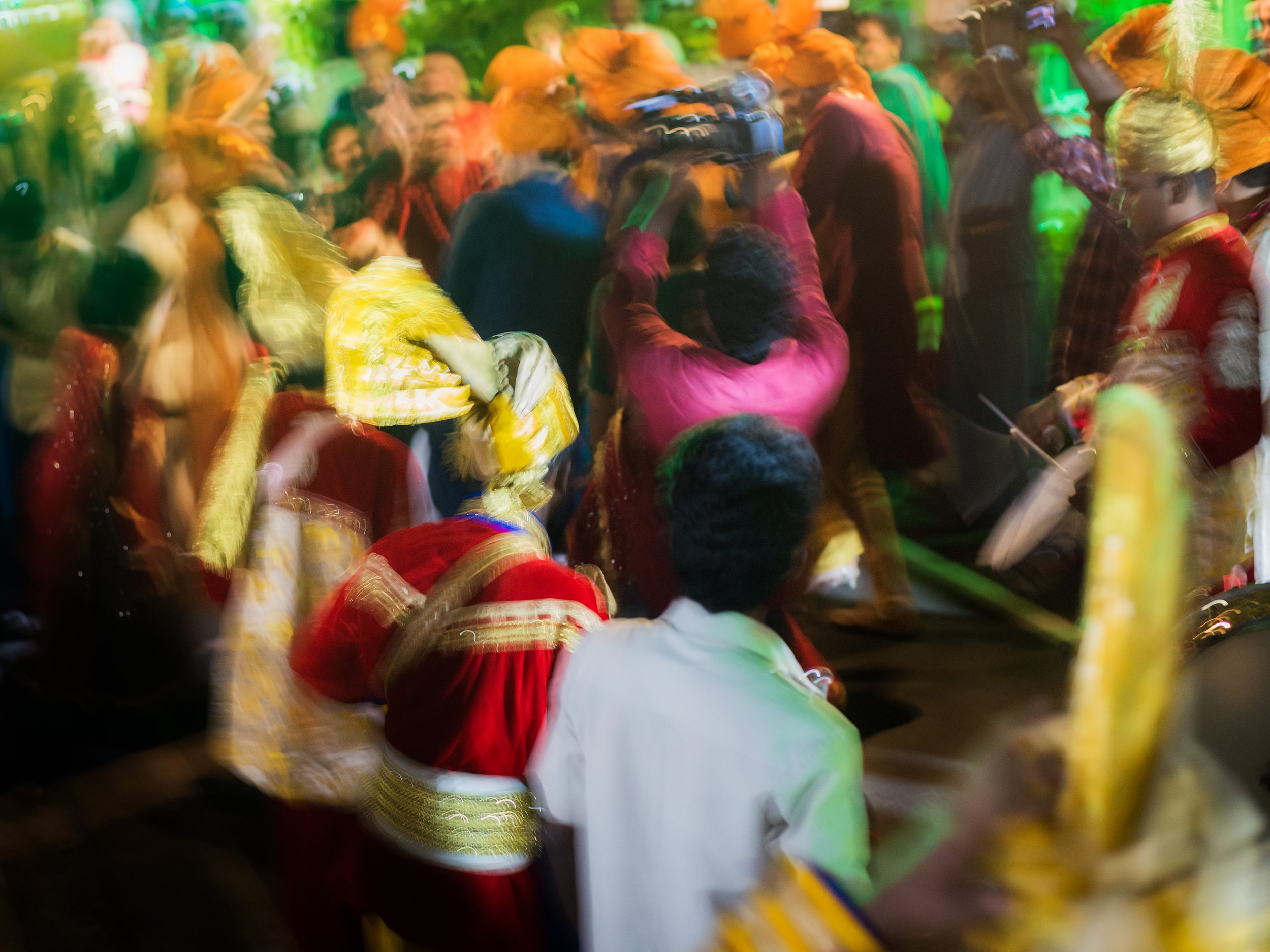 A crowded dance floor with people in colorful traditional Indian attire, including turbans and sarees, illuminated by vibrant multicolored lights.