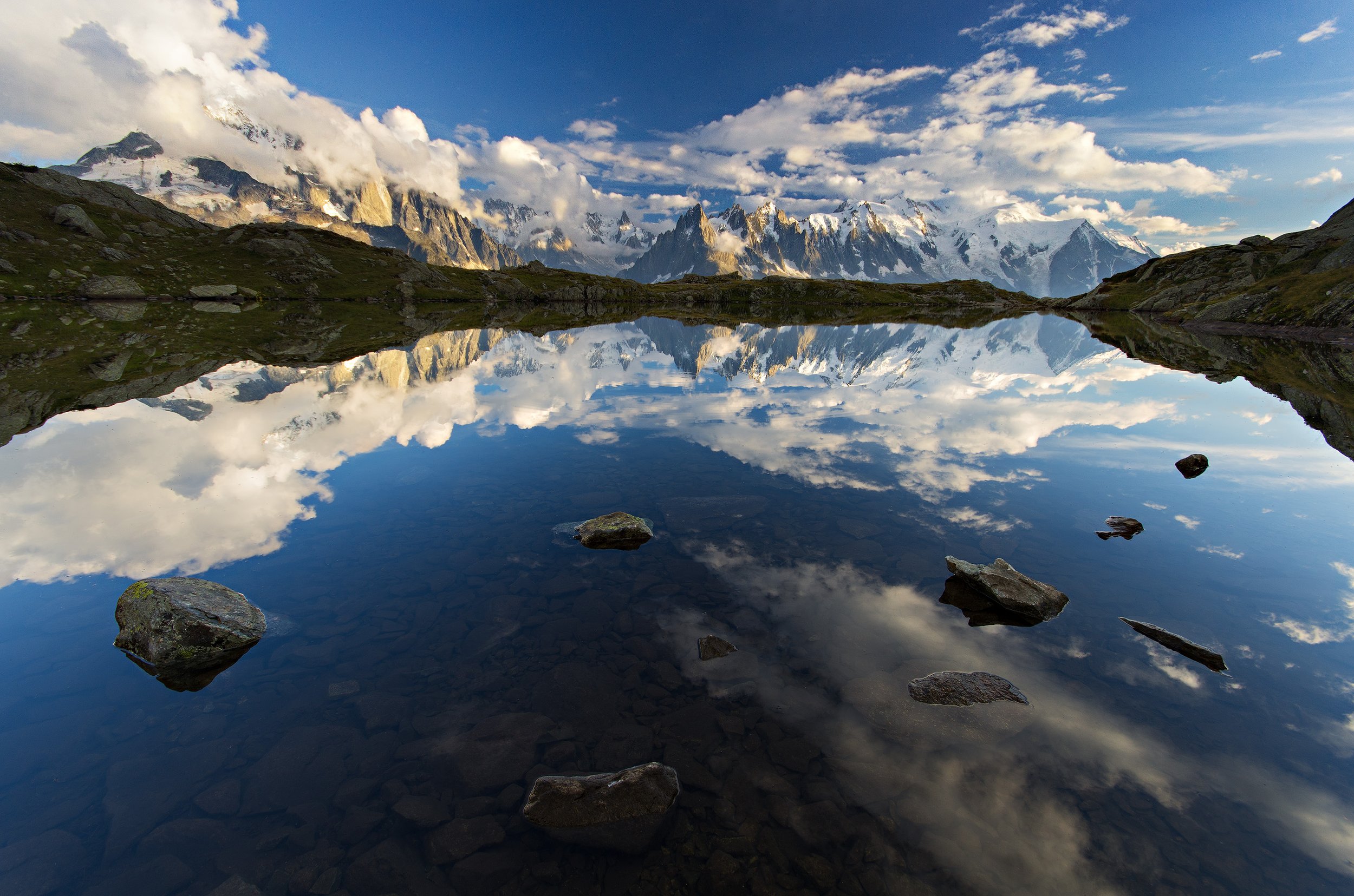 Snow-capped mountains reflected in a calm mountain lake with rocks in the foreground and a partly cloudy sky.
