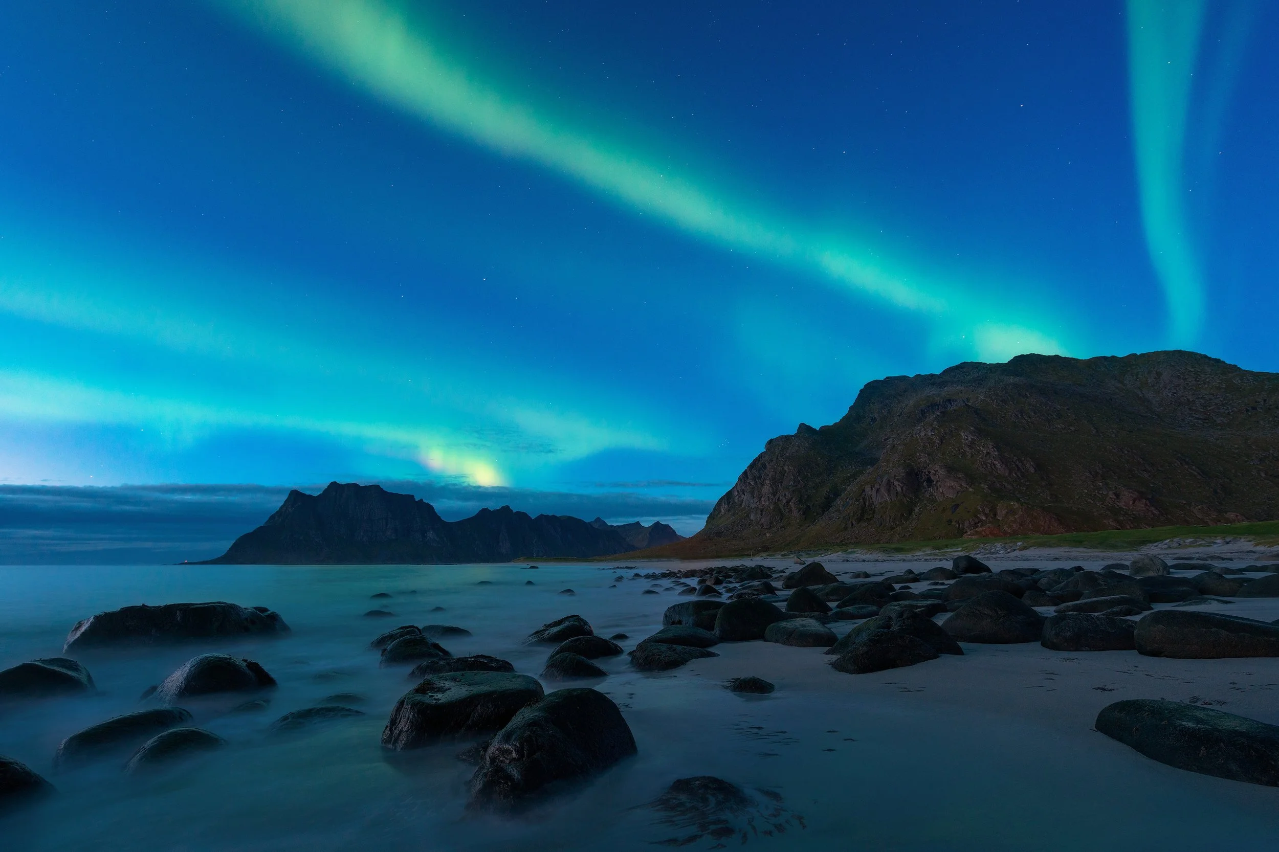 Northern lights over a rocky shoreline with mountains in the distance at night.
