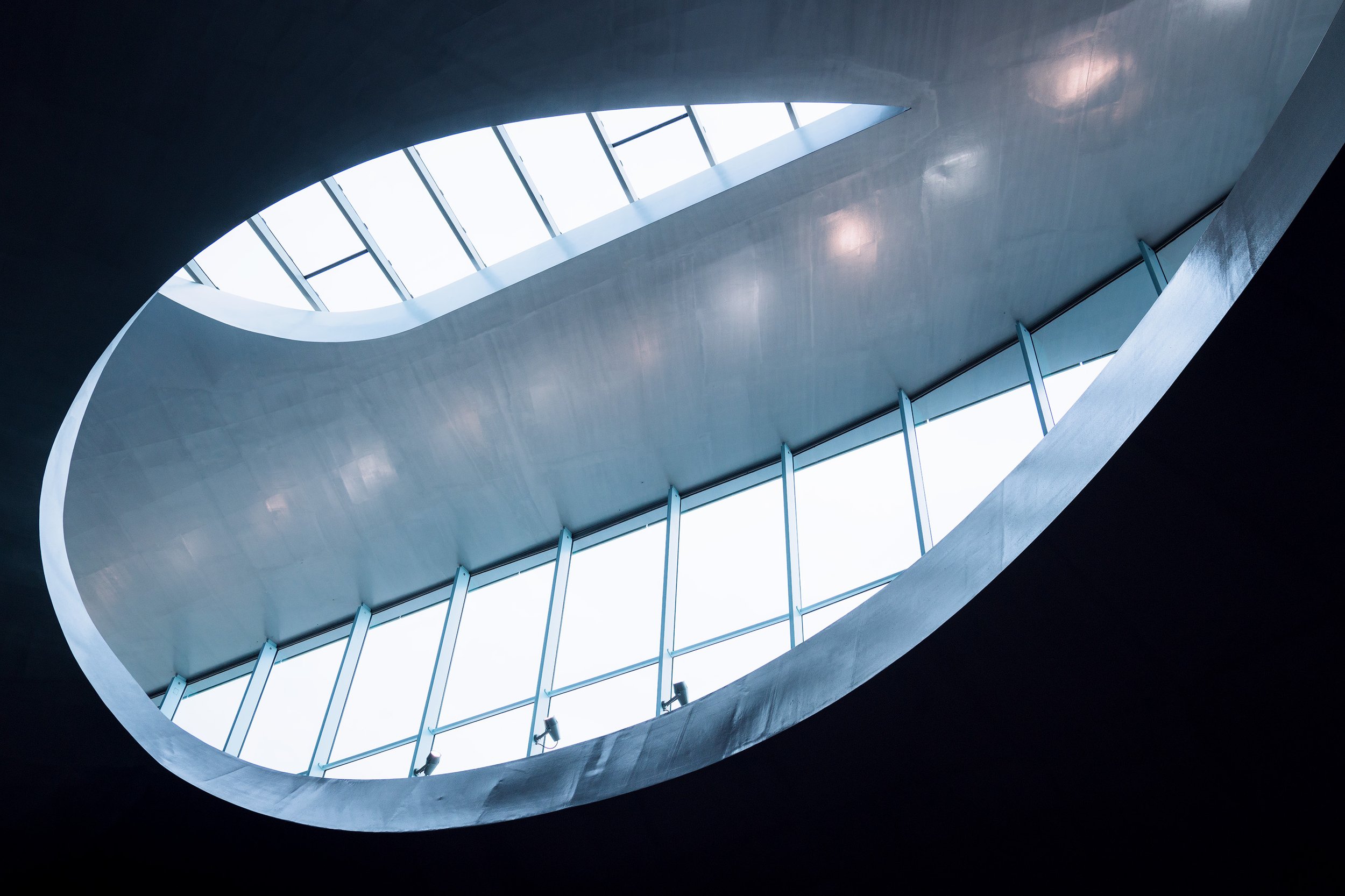 Interior view of a modern building ceiling with an oval-shaped skylight and large windows, showcasing sleek metal and glass architecture.