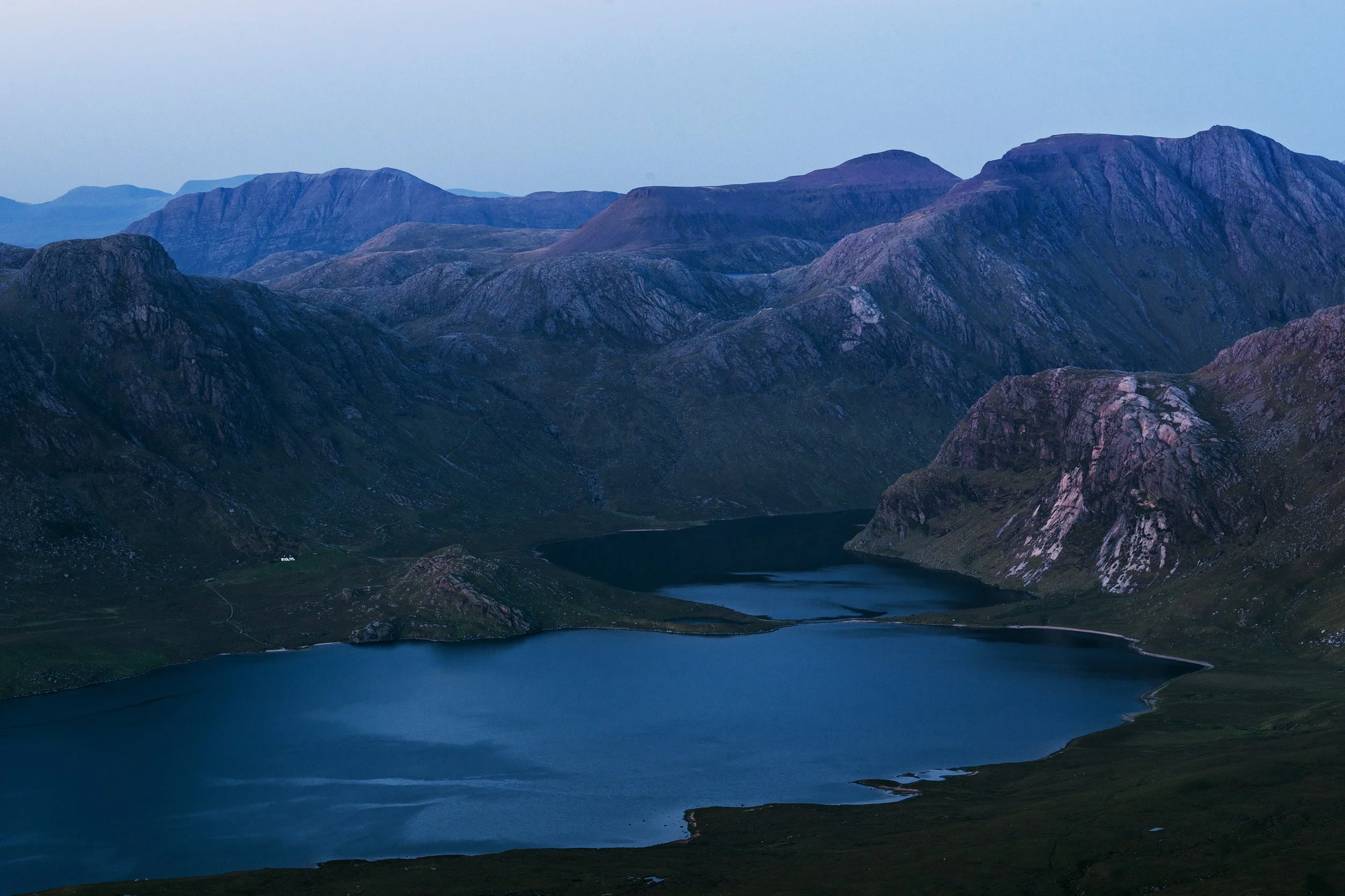 A scenic mountain landscape featuring a large blue lake surrounded by rugged, rocky mountains at dusk.
