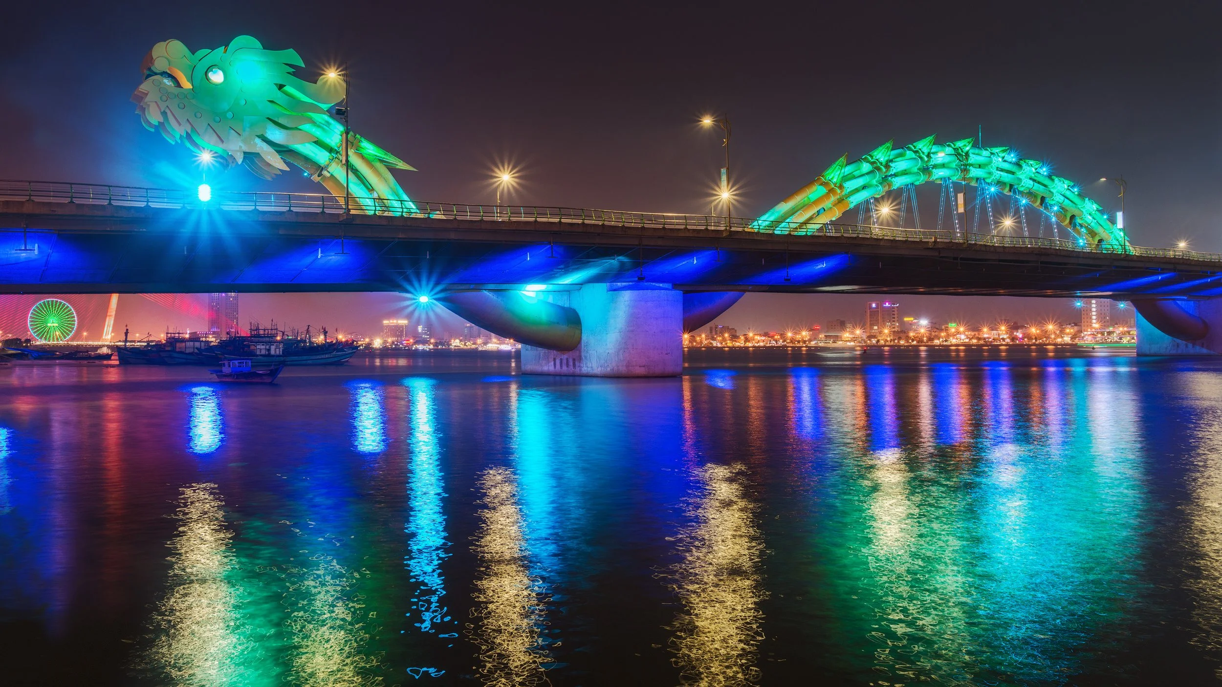 Night view of a bridge with a dragon-shaped sculpture illuminated in green and blue lights, reflecting on the water below with city lights in the background.