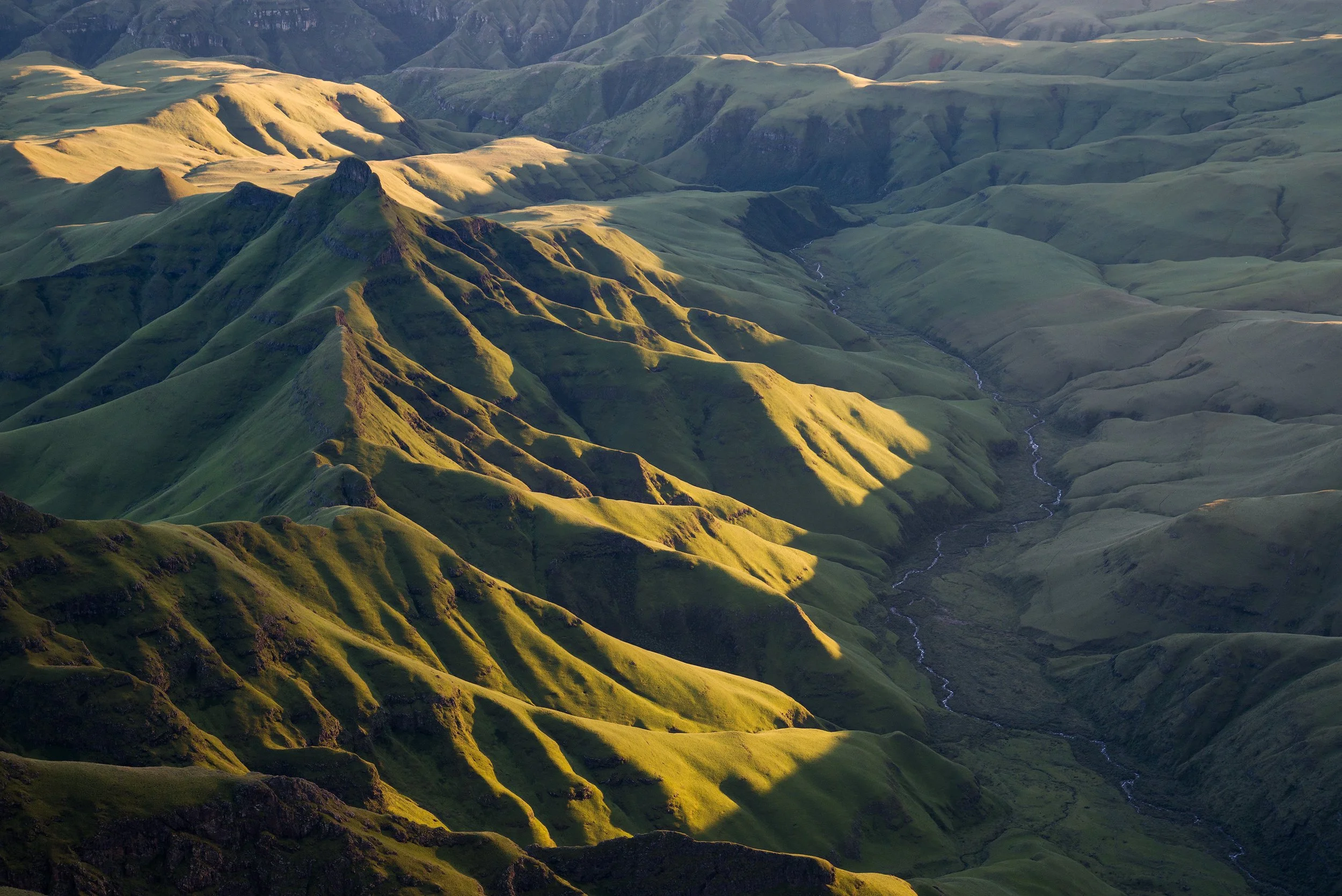 Sunlit green mountains with deep valleys and a winding stream. Drakensberg, South Africa.