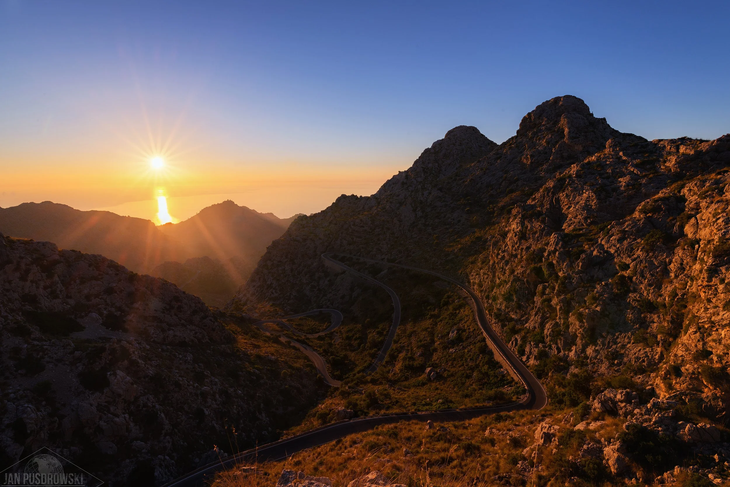 Sunset over Mallorcean mountain landscape with a winding road and rocky peaks.