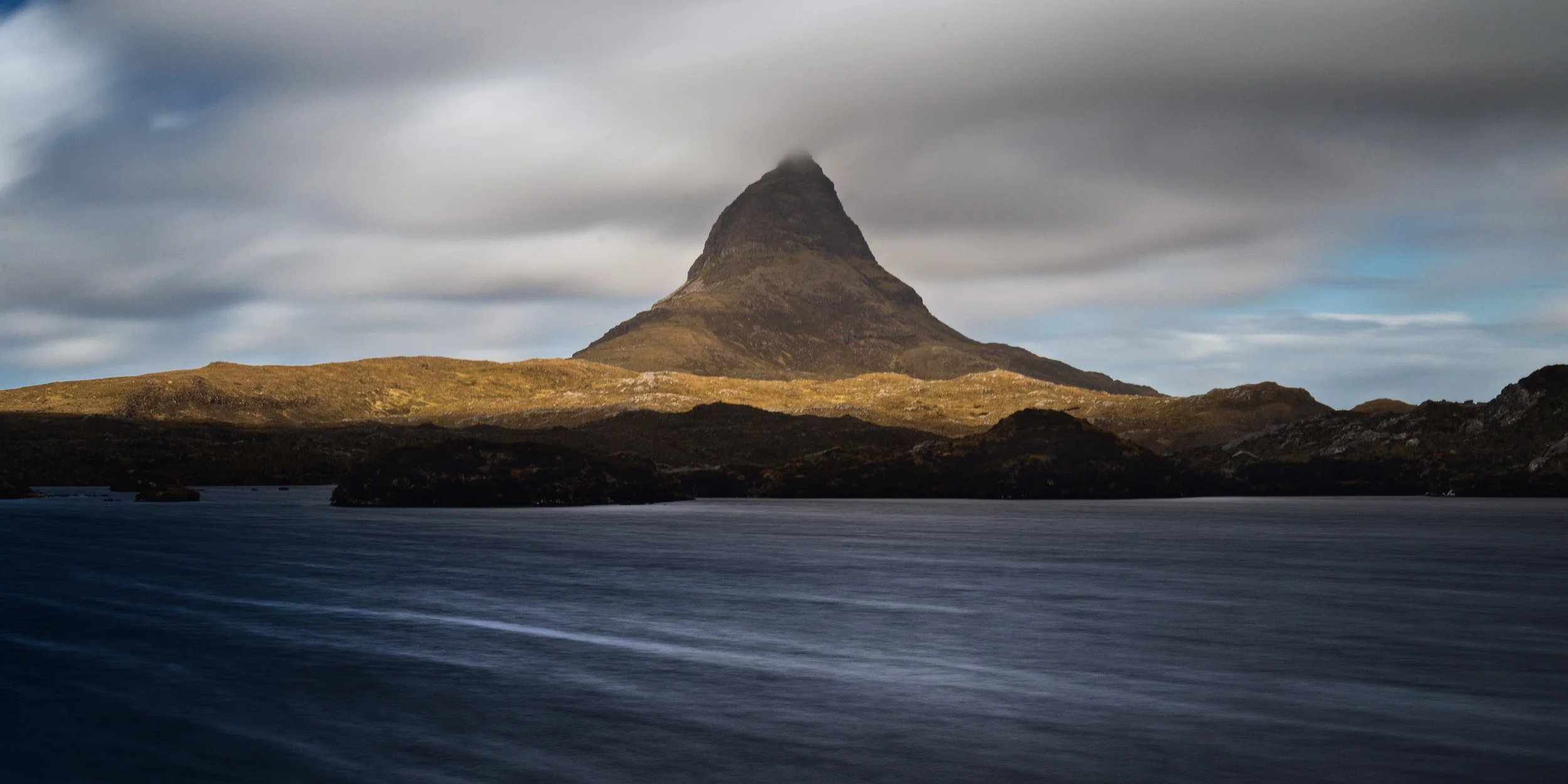 Mountain with a pointed peak partially obscured by clouds, overlooking a body of water with a dark, rocky shoreline under cloudy skies.
