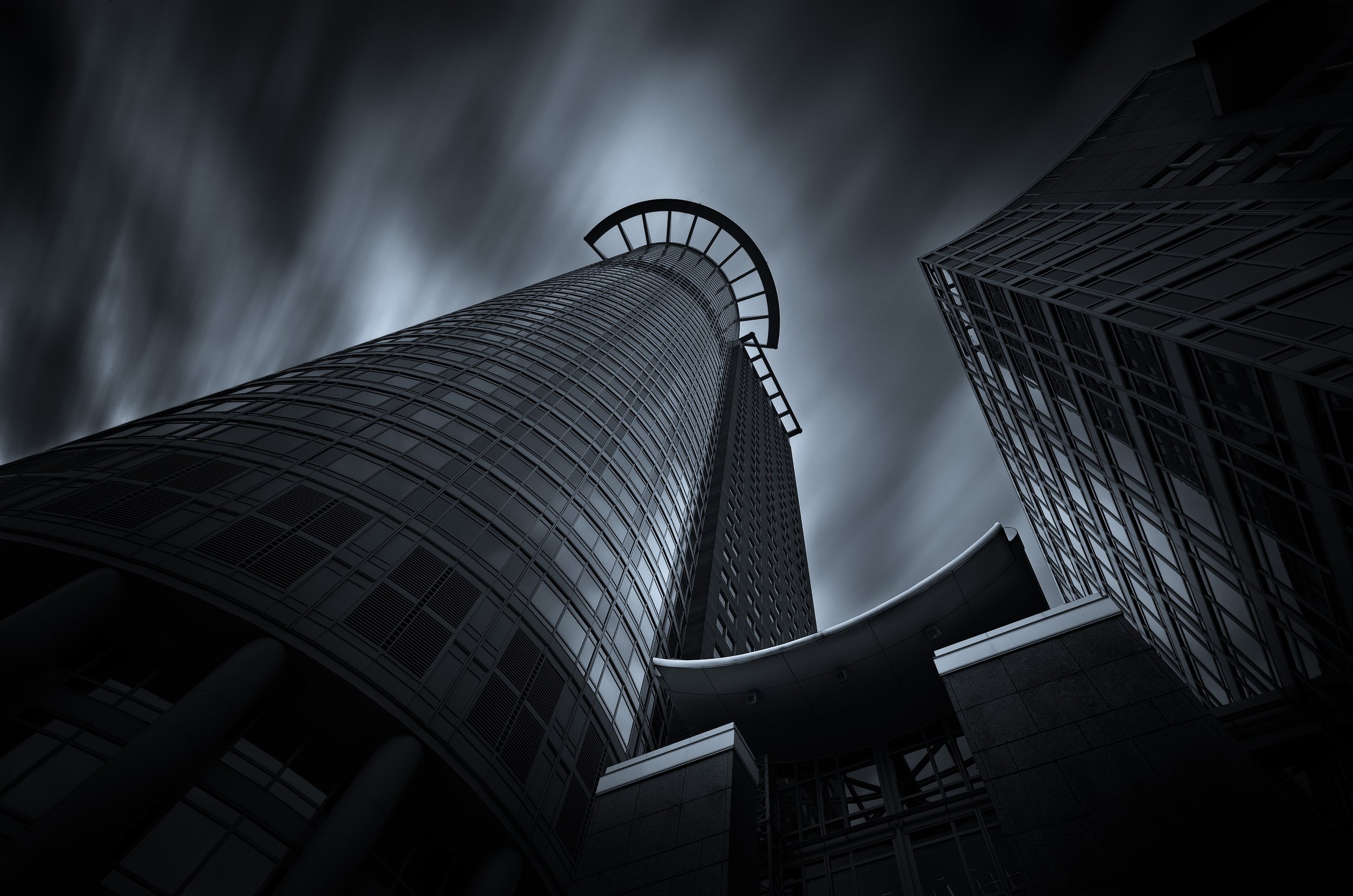 Looking up at a tall modern skyscraper with a circular observation deck near the top, dark clouds in the sky.