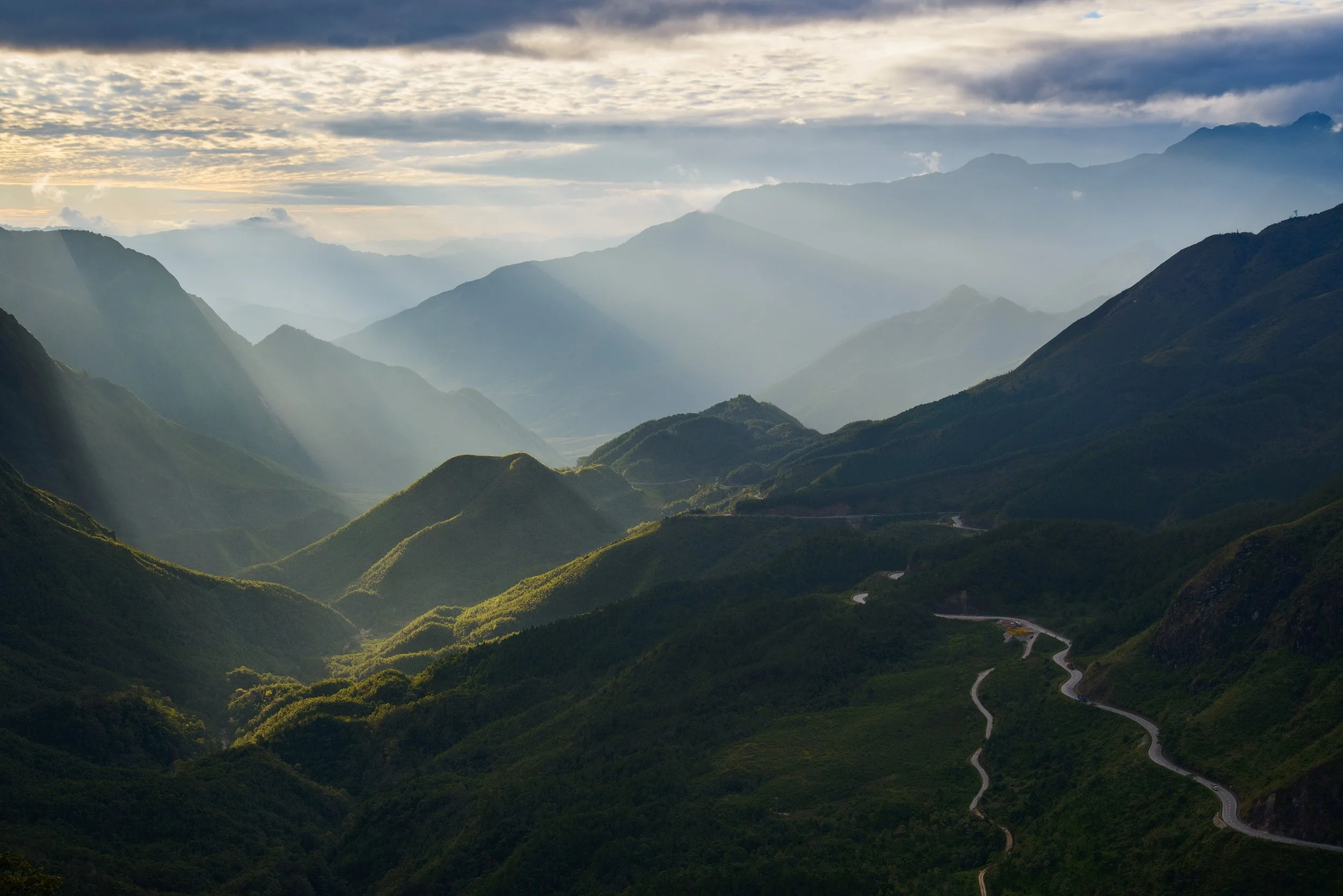 Sunlight shining through clouds over a mountain range with lush green ridges and a winding road.