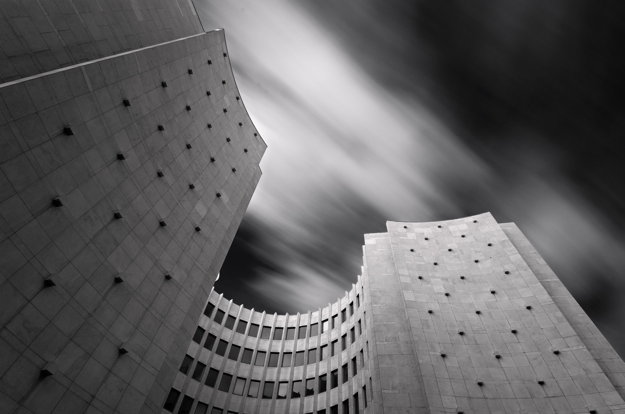 Black and white photo of modern high-rise buildings from a low angle, with clouds moving quickly in the sky.