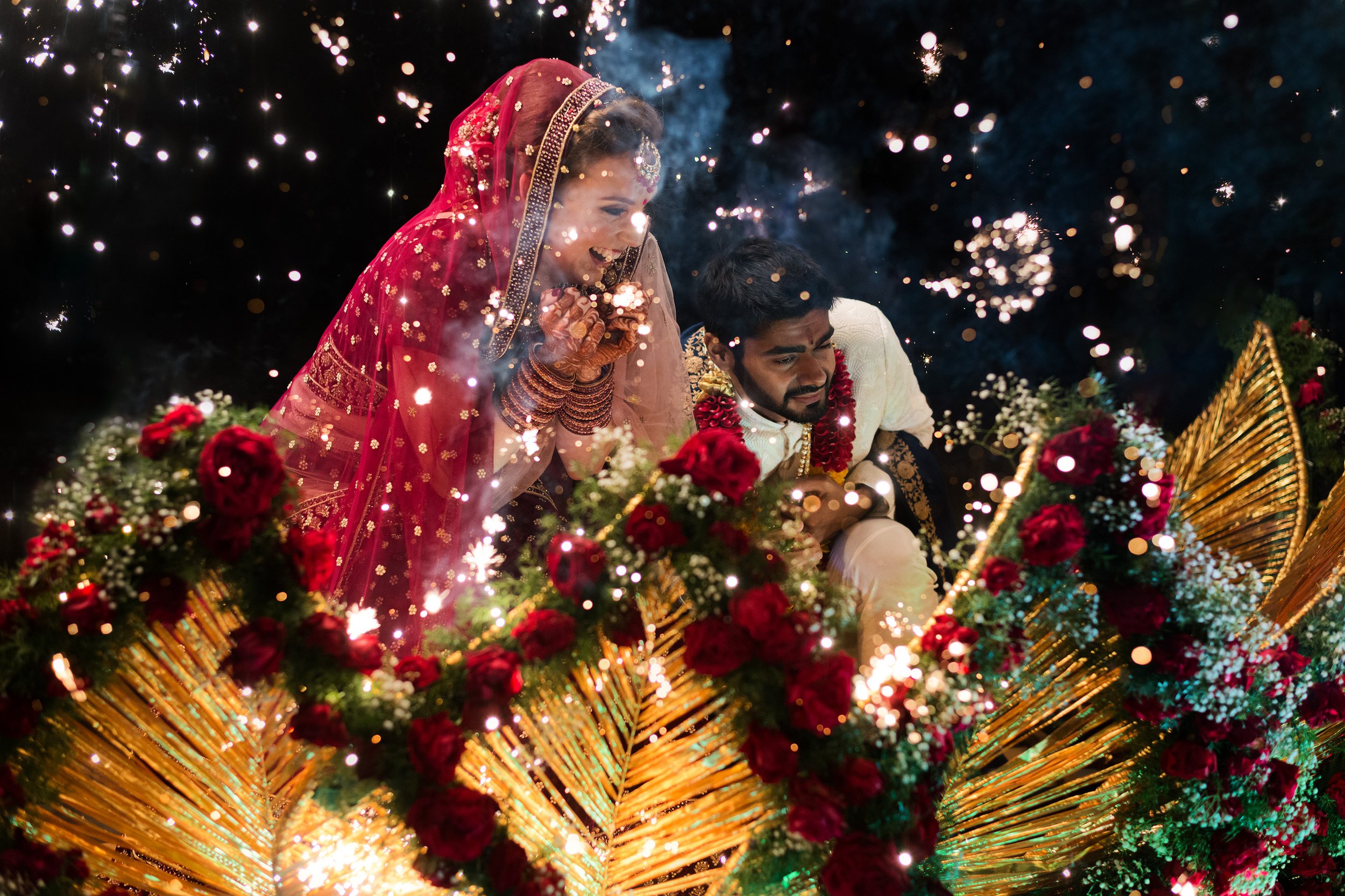Indian bride and groom celebrating during a wedding with sparks, flowers, and gold decorations.
