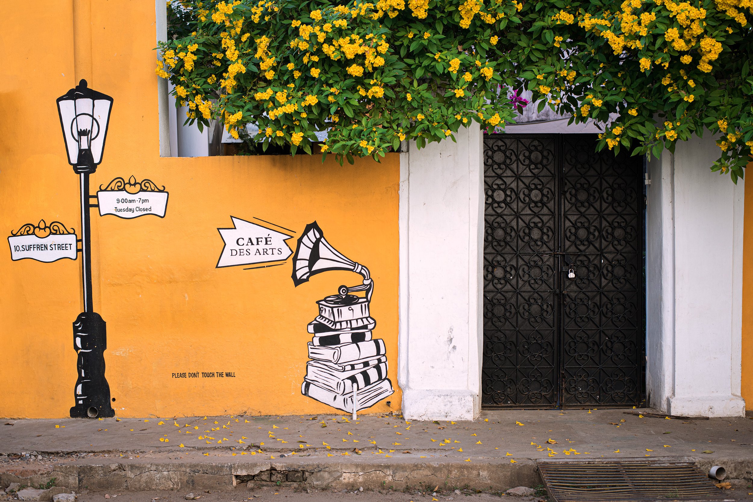 A yellow wall with artistic black and white mural of a street lamp, a banner with address '10 Sufferen Street', and a sign with business hours. The mural features a vintage gramophone atop a stack of books. Flowers and greenery are visible above the 