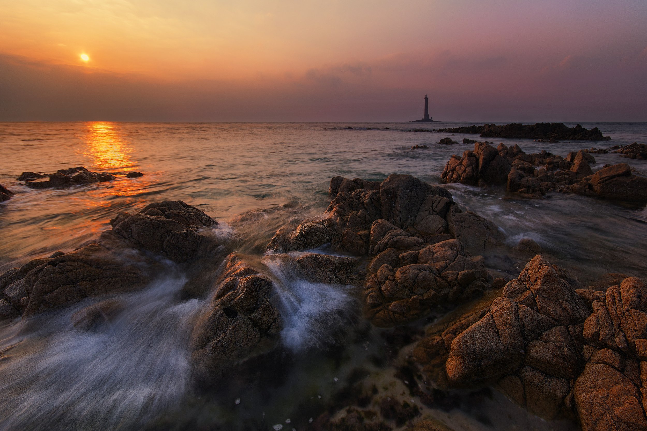 Sunset over the ocean with waves crashing over rocks, lighthouse in the distance, sky with orange, pink, and purple hues.