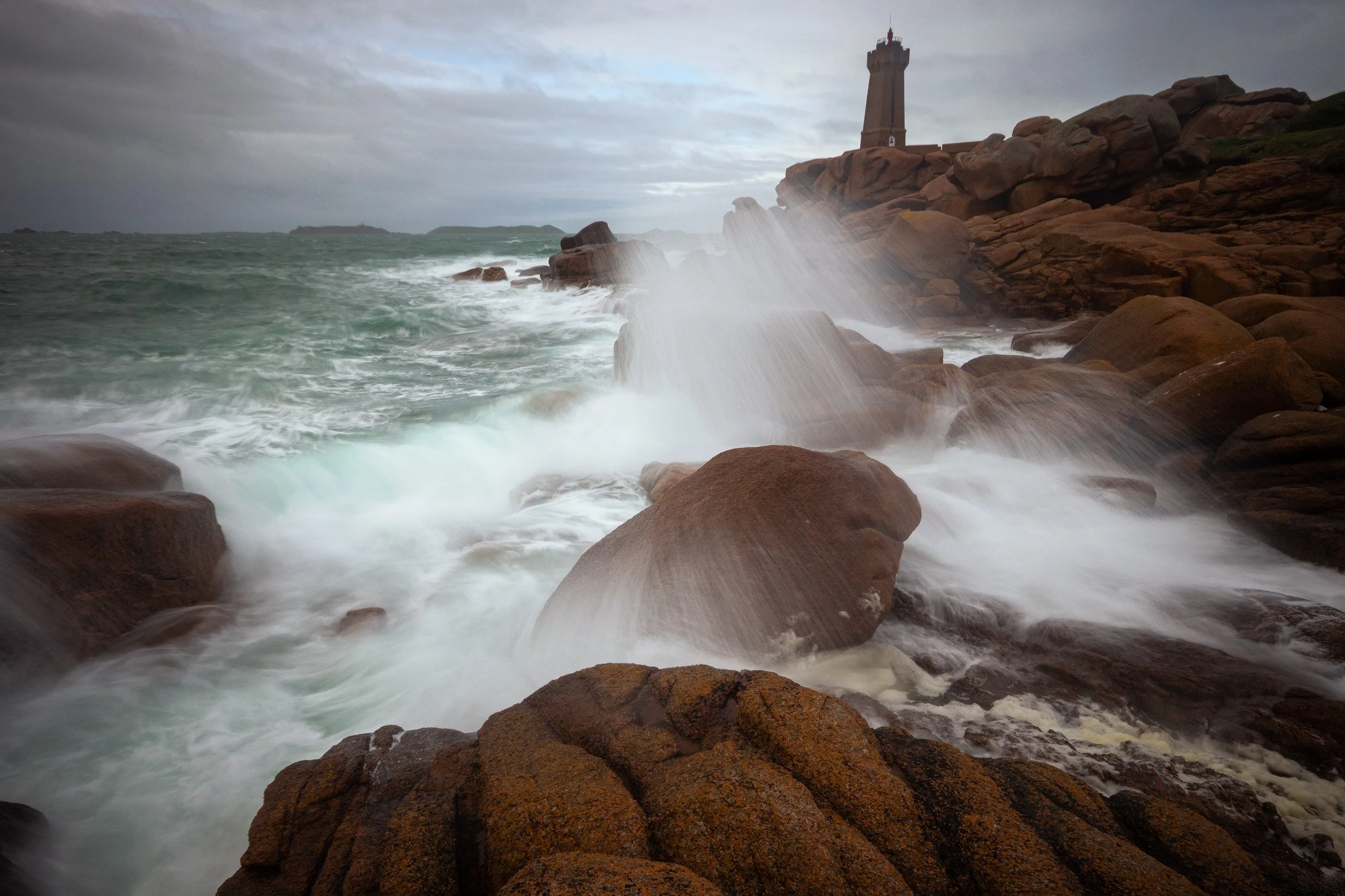 Waves crashing against rocky shoreline with a lighthouse on a cliff in the background during overcast weather.