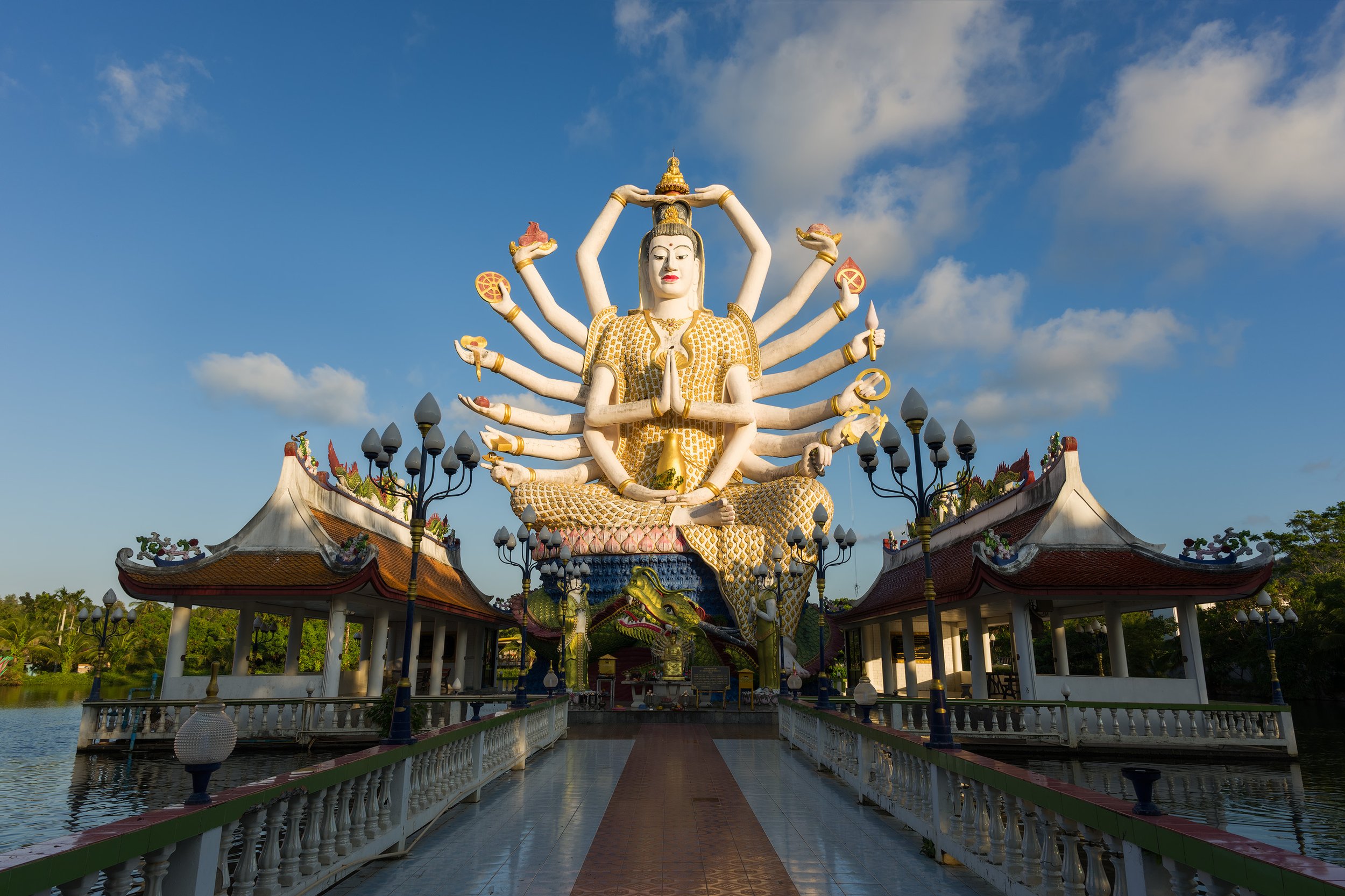 Large multi-armed Hindu goddess statue with a serene expression, seated on a platform with a dragon motif at the base, surrounded by pavilions, lanterns, and water, under a partly cloudy sky. Koh Samui, Thailand.