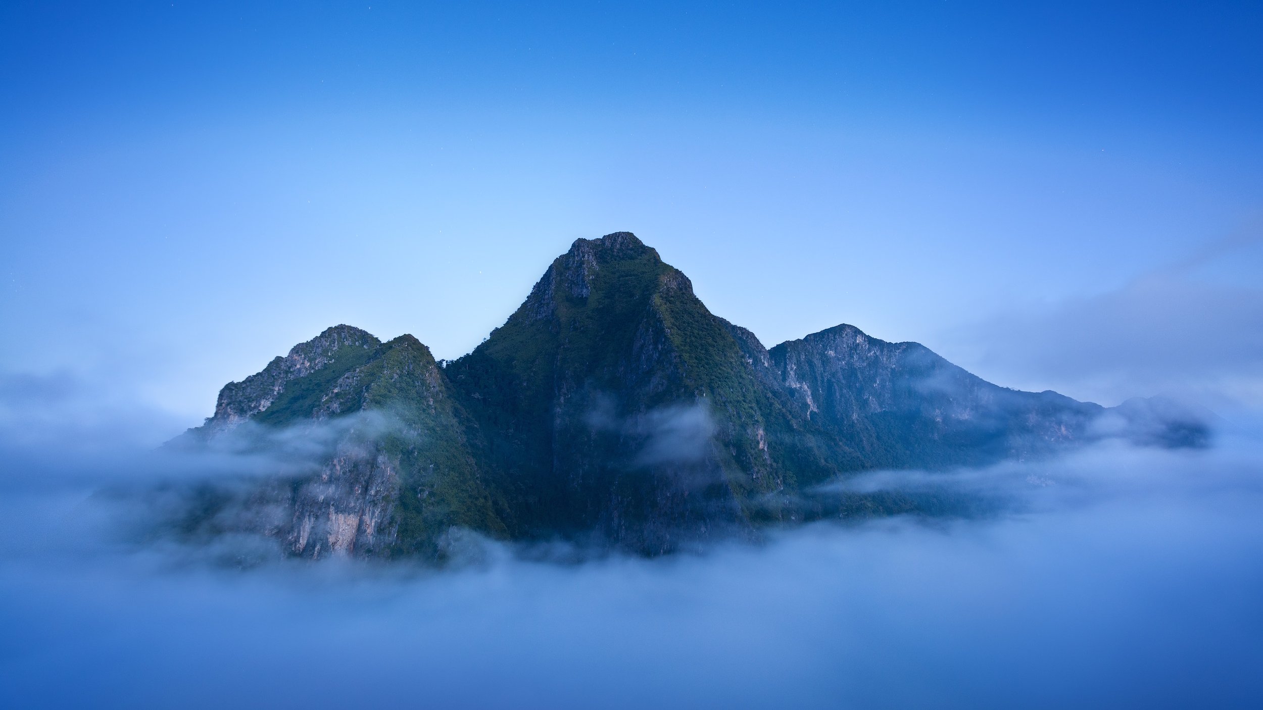 Mountain landscape with peaks covered in green vegetation, surrounded by mist and clouds during twilight sky. Laos rugged peaks.