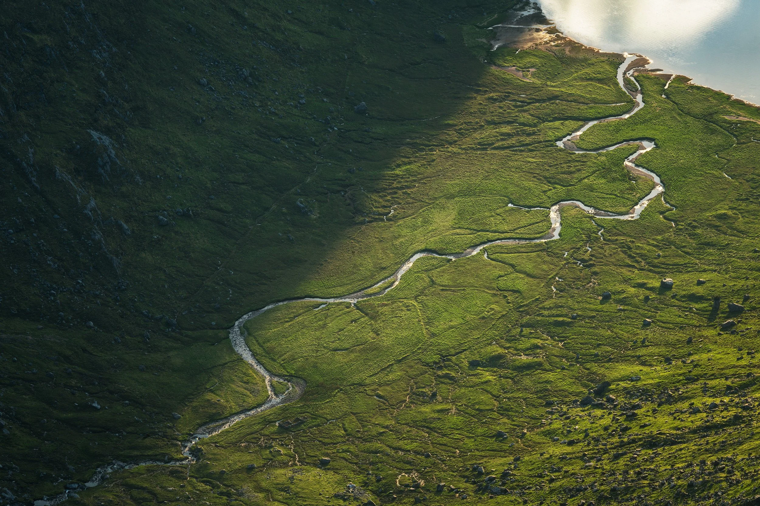 A winding stream flowing through a green valley with hills and a body of water in the background.