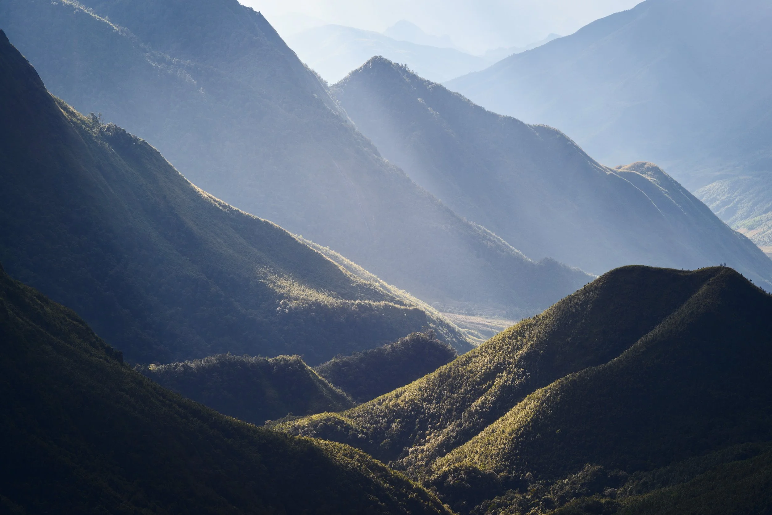 Multiple green mountains with varying shades, layered and covered in greenery, under a light sky with some mist or fog. Sapa, Viet Nam, China.